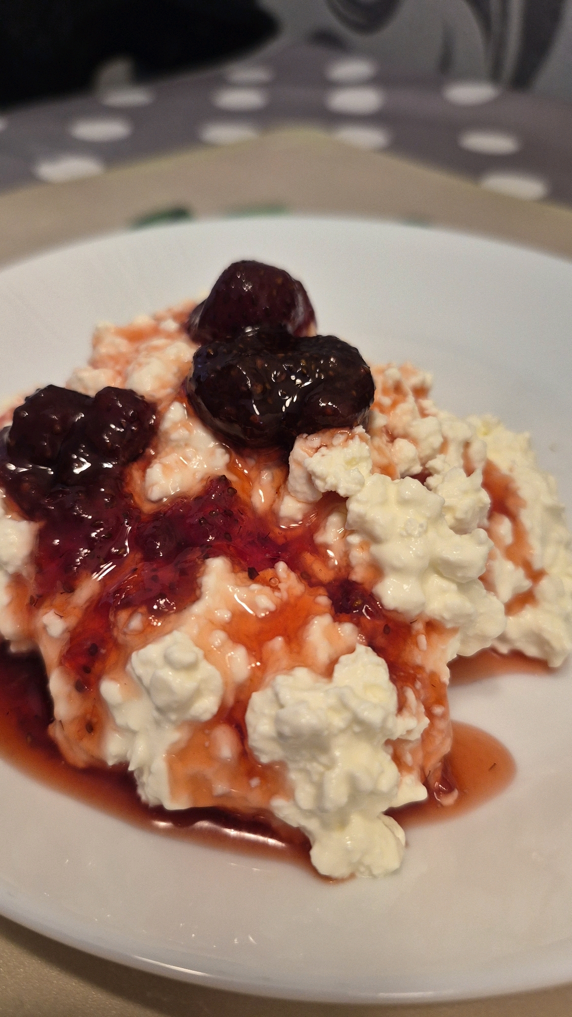 This is a close-up, eye-level, indoor shot of a white plate of cottage cheese topped with jam and whole strawberries. The cottage cheese is a lumpy, white texture. The jam is a bright red color with visible seeds, and it is drizzled over the cottage cheese. Three whole, dark red strawberries are placed on top of the dish. The plate is on a surface that appears to be a tablecloth with a dotted pattern in grey and white. The background is out of focus, showing a hint of a dark object and the textured tablecloth. There are no people visible in the image. The setting appears to be a home environment, likely a kitchen or dining area in Krauja, Latvia.