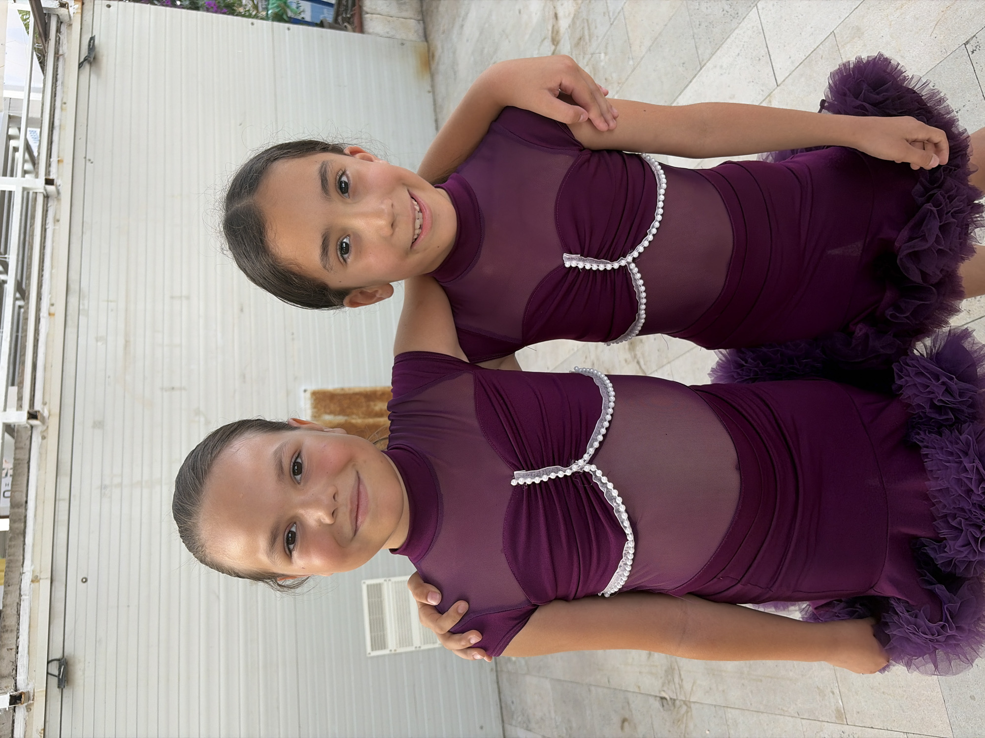 Two young girls, likely sisters, pose for a photo in matching purple, ruffled dance costumes. The girls have dark hair and are smiling at the camera. The costume features sheer sleeves and a sheer neckline with pearl detailing along the bust line. The skirt of the costume is short and has layers of ruffles.
The setting appears to be outdoors, possibly a patio or walkway, with a textured stone floor and a corrugated metal wall in the background. There is also a glimpse of some greenery and a white metal structure that could be a fence or part of a building. The lighting suggests it is daytime. The girls are positioned close together, with one girl slightly in front of the other, and their arms are around each other. Their happy expressions and coordinated outfits suggest they are participating in a dance event or performance.
No visible text is present in the image. The location context is Budva, Montenegro.