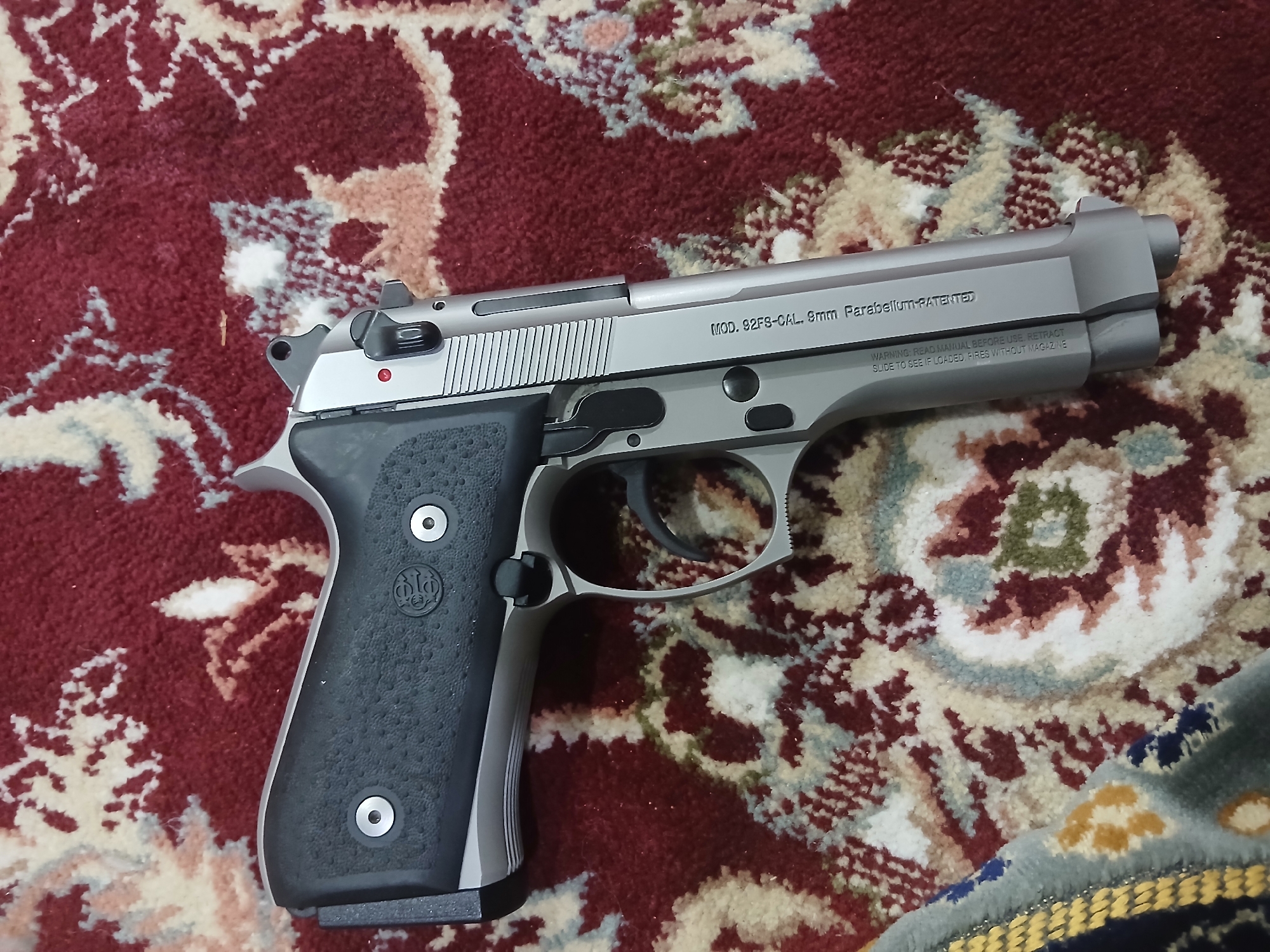 A silver Beretta 92FS pistol lies on a patterned rug. The pistol is oriented with the barrel pointing towards the upper right corner of the image. The black grip features the Beretta logo. Text on the side of the slide reads "MOD. 92FS-CAL. 9mm Parabellum-PATENTED" and a warning about reading the manual before use.