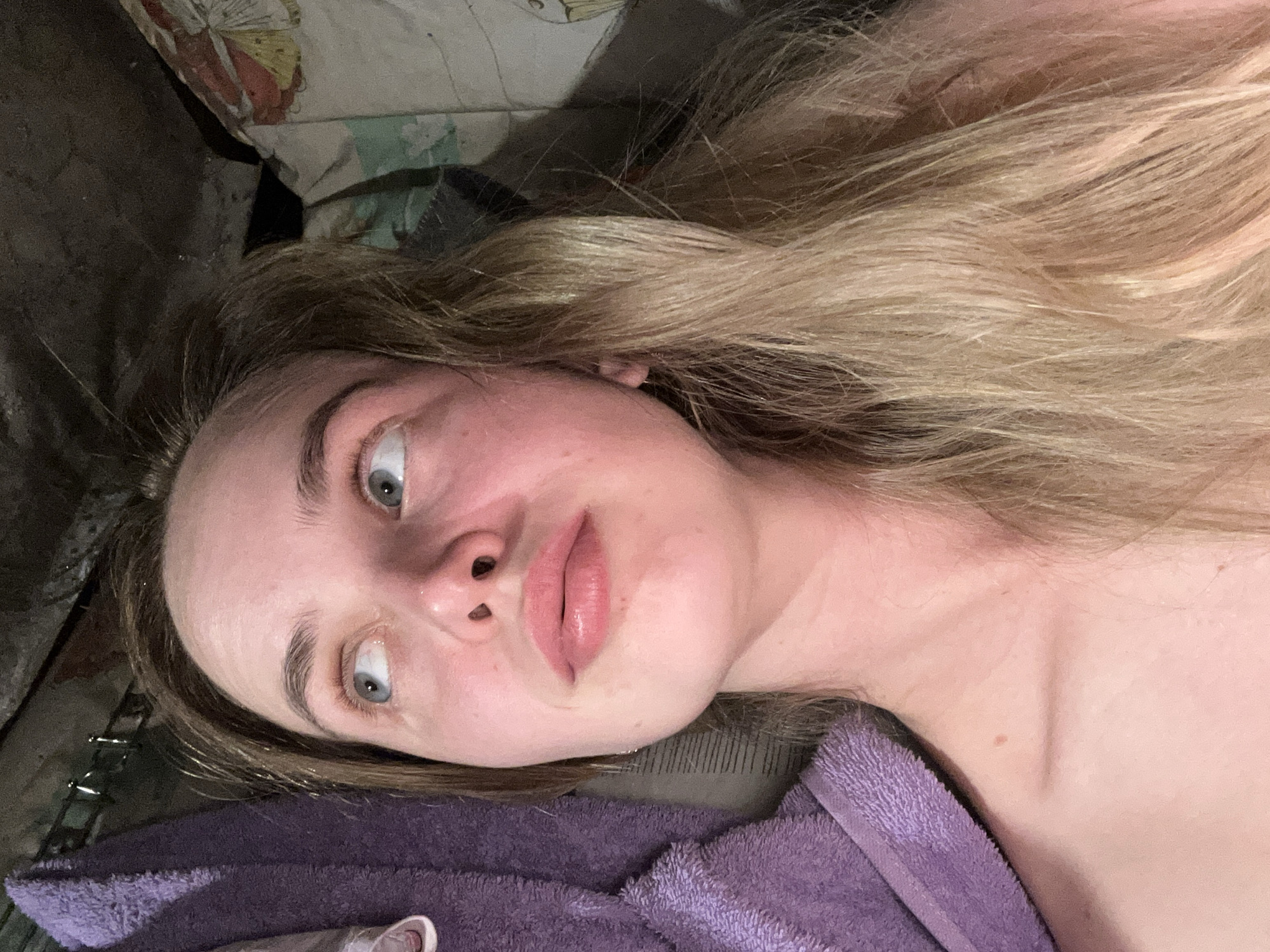 A close-up shot features a young woman with blonde hair, looking upwards and to her left. She is lying down, and a portion of a purple towel or robe is visible on the left side of the frame. The background is dark and appears to be an indoor setting with some indistinct patterns on what might be bedding or upholstery on the right. The lighting is dim, casting shadows. The location is identified as Азинский, Russia.