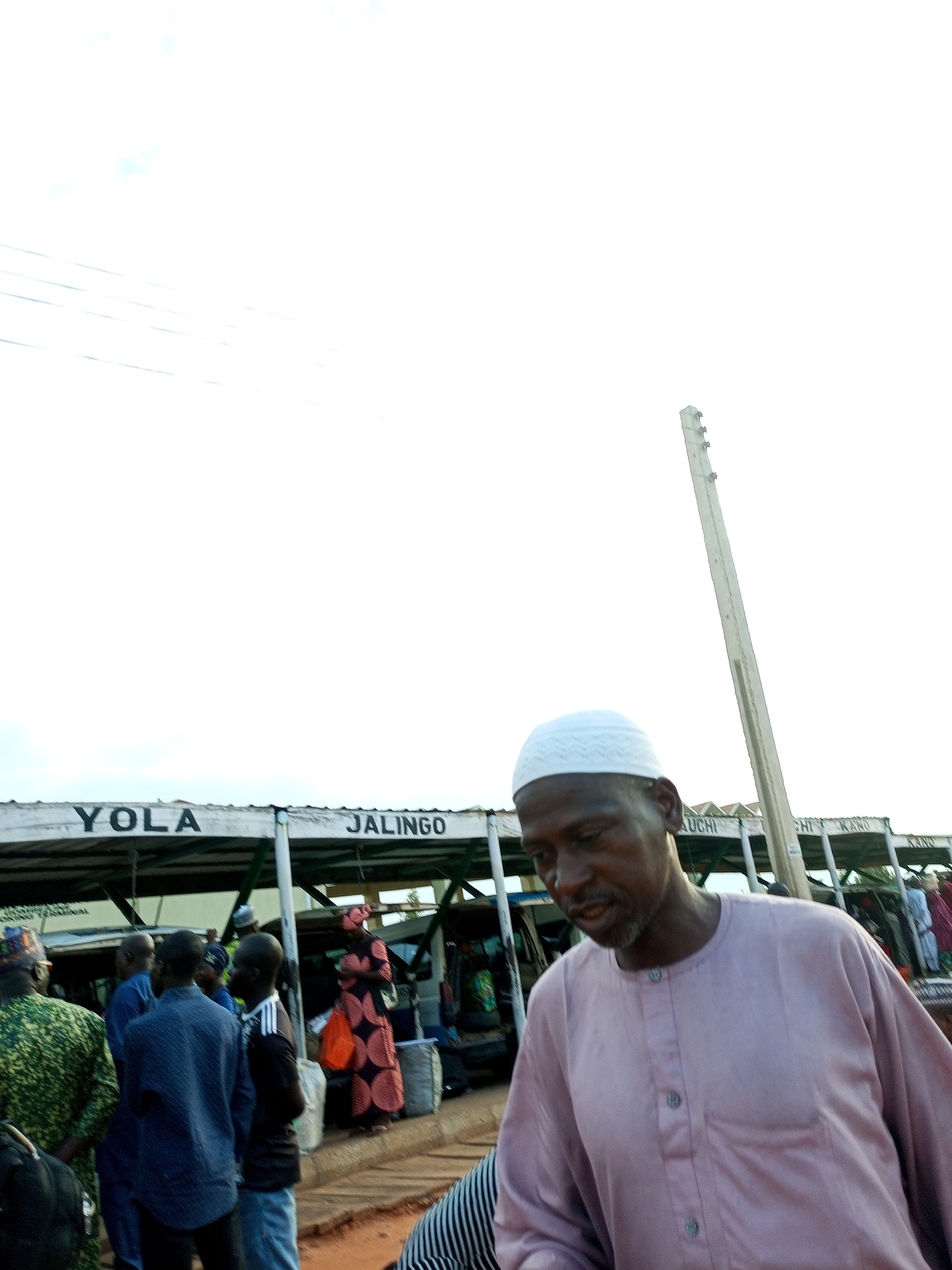 The image shows an outdoor scene at what appears to be a bus station or terminal, likely in Gombe, Nigeria. In the foreground, a man wearing a light purple tunic and a white kufi cap is looking down and to his left. Behind him, a white pole stands vertically, angled towards the right. Further back, a covered structure with signs is visible. The signs read "YOLA" and "JALINGO," indicating destinations or routes. Several other people are present, some standing and conversing, while others appear to be passengers with luggage. One woman in the background wears a patterned dress and carries an orange bag. The overall environment suggests a bustling transportation hub.