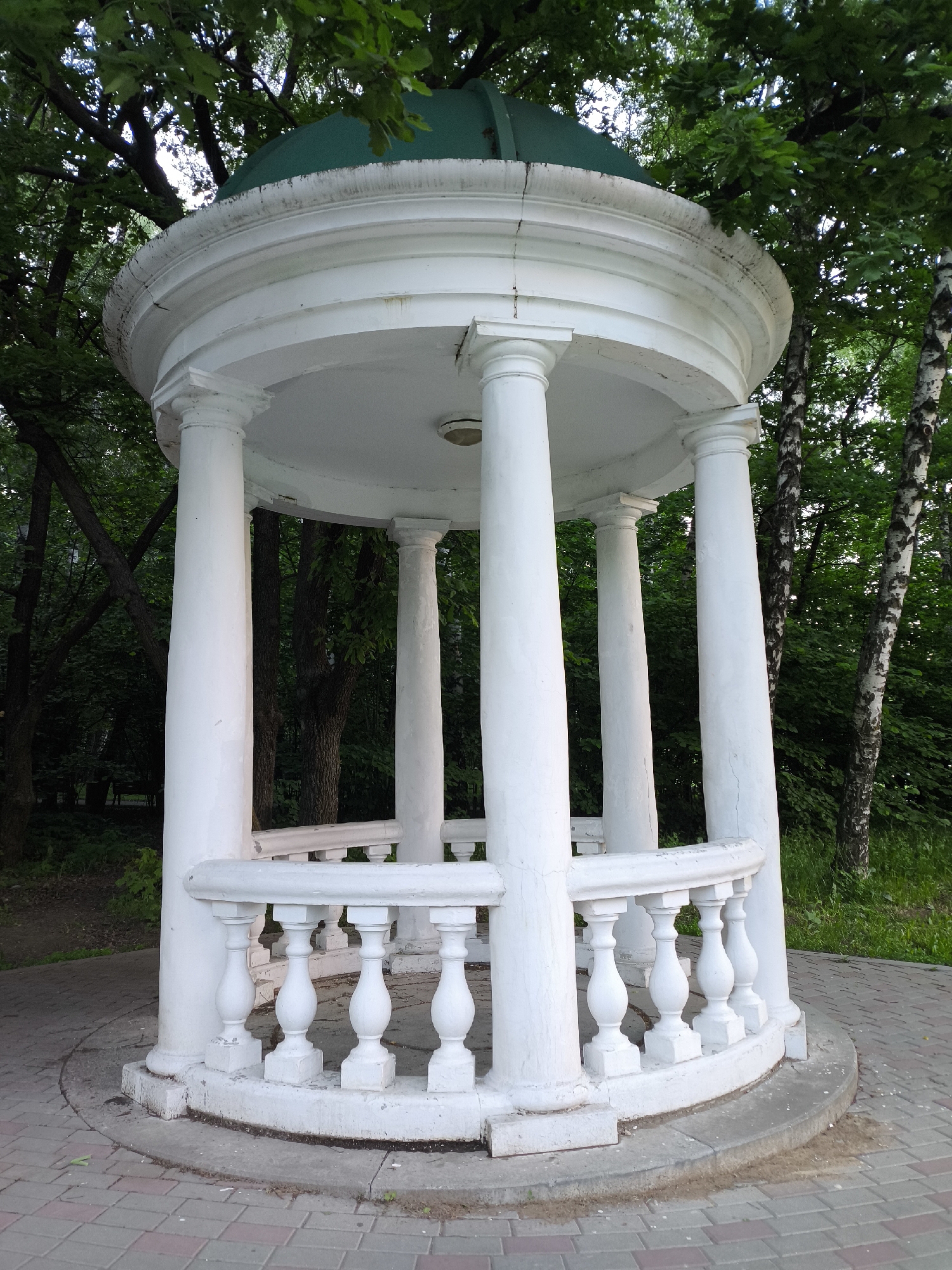 The image captures a white, classical-style gazebo set within a lush green park or garden environment. The gazebo is the central subject, featuring a circular design with a balustrade railing composed of white balusters around its base. Several robust, unfluted white columns support a flat white ceiling and a distinct green, slightly domed roof.
The structure shows signs of age and weathering, with visible dirt and discoloration on the green roof and the upper architectural moldings, as well as minor cracks. The base of the gazebo rests on a paved circular area made from interlocking red and grey rectangular bricks.
Surrounding the gazebo is dense green foliage, including numerous trees and bushes, forming a verdant backdrop. Notably, on the right side, the distinctive white and black bark of birch trees is visible. The lighting suggests it is daytime, possibly morning or late afternoon, with soft, diffused light filtering through the tree canopy. No people are present, and no specific activity is taking place. There is no visible text in the image. The scene conveys a sense of peaceful tranquility, showcasing an aged architectural feature nestled in a natural, serene setting.