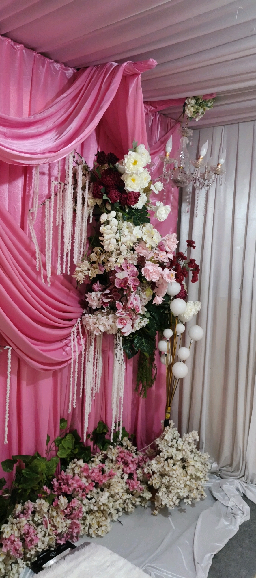 This is a vertical, eye-level shot of a decorative floral arrangement in Muaro Jambi, Indonesia. The arrangement is positioned in front of a pink draped backdrop and a white curtain. The flowers are a mix of white, cream, pink, and deep red, with various types of blooms and greenery. Dangling white tassels hang from the top of the pink drapery. A crystal chandelier is visible above the arrangement. To the right of the floral display, a gold-colored stand with white spherical accents is partially visible. The foreground shows a carpet of pink and white artificial flowers, with some dark green artificial leaves. A white fluffy rug and a black object are visible in the lower left corner.