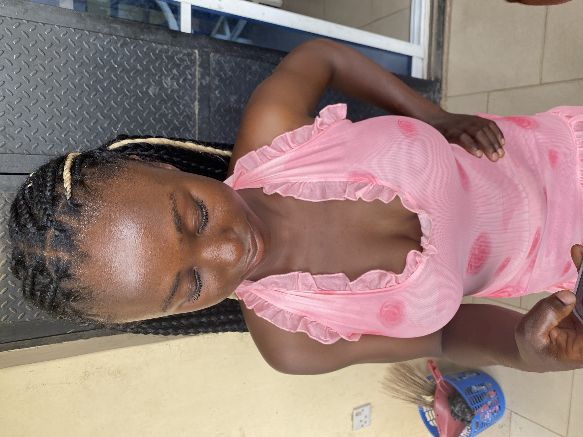 A dark-skinned woman is depicted lying down or reclining in a relaxed posture, with her eyes closed, suggesting rest or serenity. She has black braided hair, interspersed with blonde strands. Her skin appears smooth, with a subtle sheen on her forehead.
She is dressed in a light pink, sheer garment, possibly a nightgown, featuring ruffled edges around the neckline and sleeves. The fabric of her clothing has faint circular patterns. Her right arm is bent with her hand resting on her abdomen, while her left hand is partially visible, holding what appears to be a mobile phone.
The setting, located in Jos, Nigeria, features a dark, textured metallic surface resembling diamond plate behind her head and upper body. The lower part of the image shows a light-colored floor, possibly tiled or concrete. On this floor, a blue plastic basket containing a broom and a dustpan is visible, alongside a white electrical outlet. A white and blue structural element, potentially part of a window or door frame, can be seen in the background near her left arm. The bright, even lighting indicates it is daytime. There is no visible text in the image.