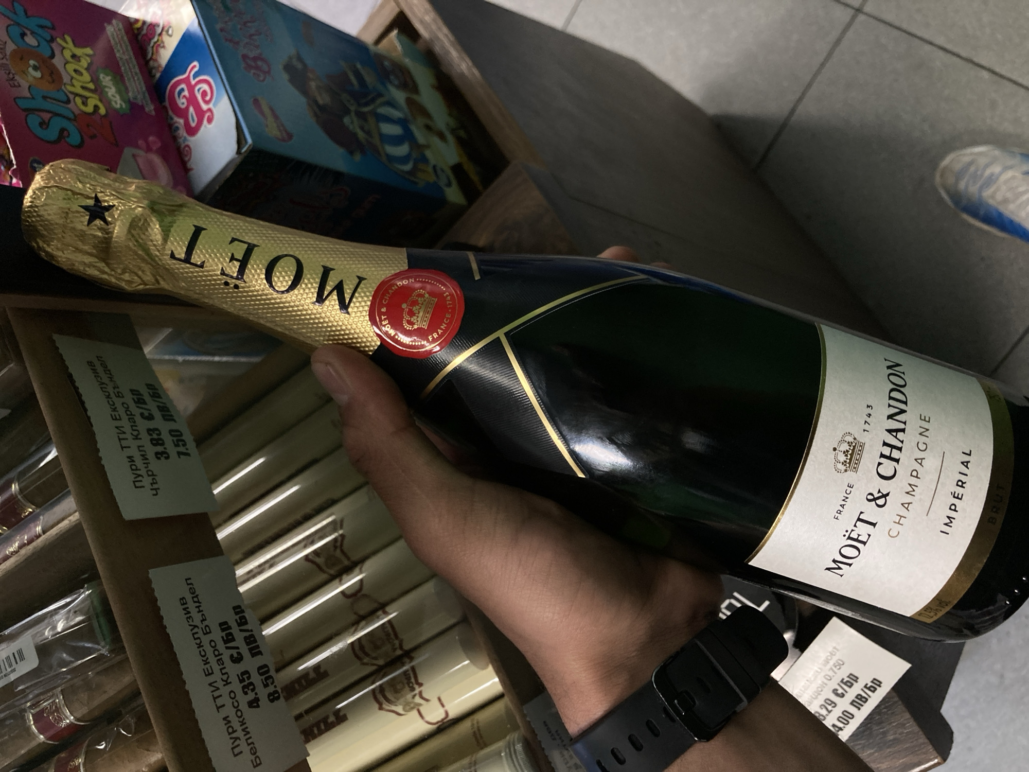 The image shows a hand holding a bottle of Moët & Chandon Impérial Brut champagne. The bottle is positioned over a shelf displaying cigars in individual plastic wraps with pricing labels in Cyrillic script. A price tag associated with the bottle indicates "Гандон 0.750" (likely a price per bottle), with prices listed in both euros (€) and Bulgarian lev (ЛВ/Бр). The background includes boxes of candy with cartoonish branding. The photograph seems to have been taken in a store, potentially in Kazichene, Bulgaria, given the pricing and language displayed on the labels. The location within the city cannot be determined solely from this image.