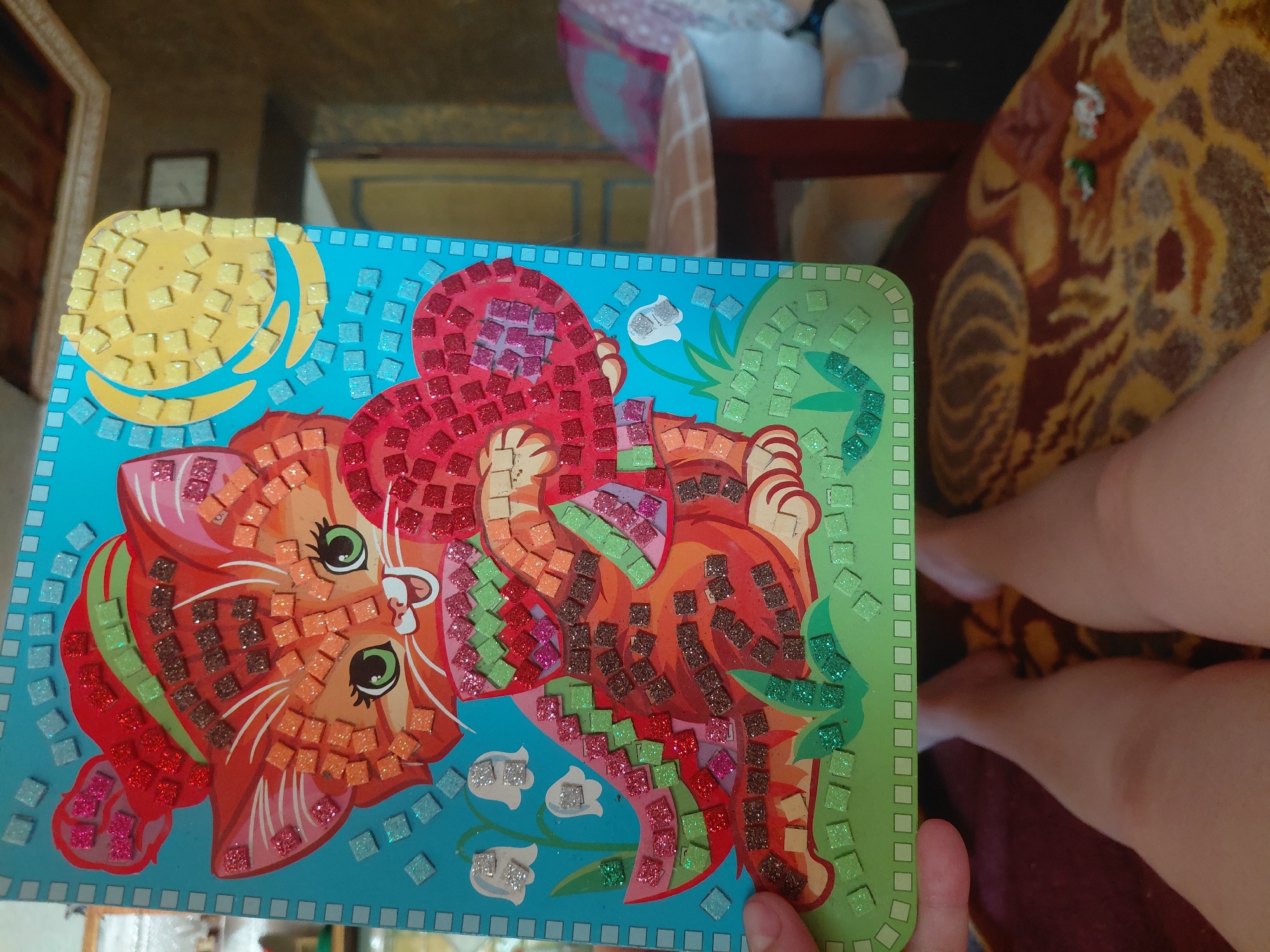 An image captures a woman's legs and a mosaic craft depicting a cat holding a heart, positioned indoors. The craft is likely a children's activity kit, characterized by its colorful glittery mosaic tiles forming the image of a ginger cat with a red hat, a green and red striped scarf, and a red heart. The background reveals an indoor setting with a patterned rug and a glimpse of furniture. The specific location of Marynivka, Ukraine, is not discernible from the visual information presented in the image.
