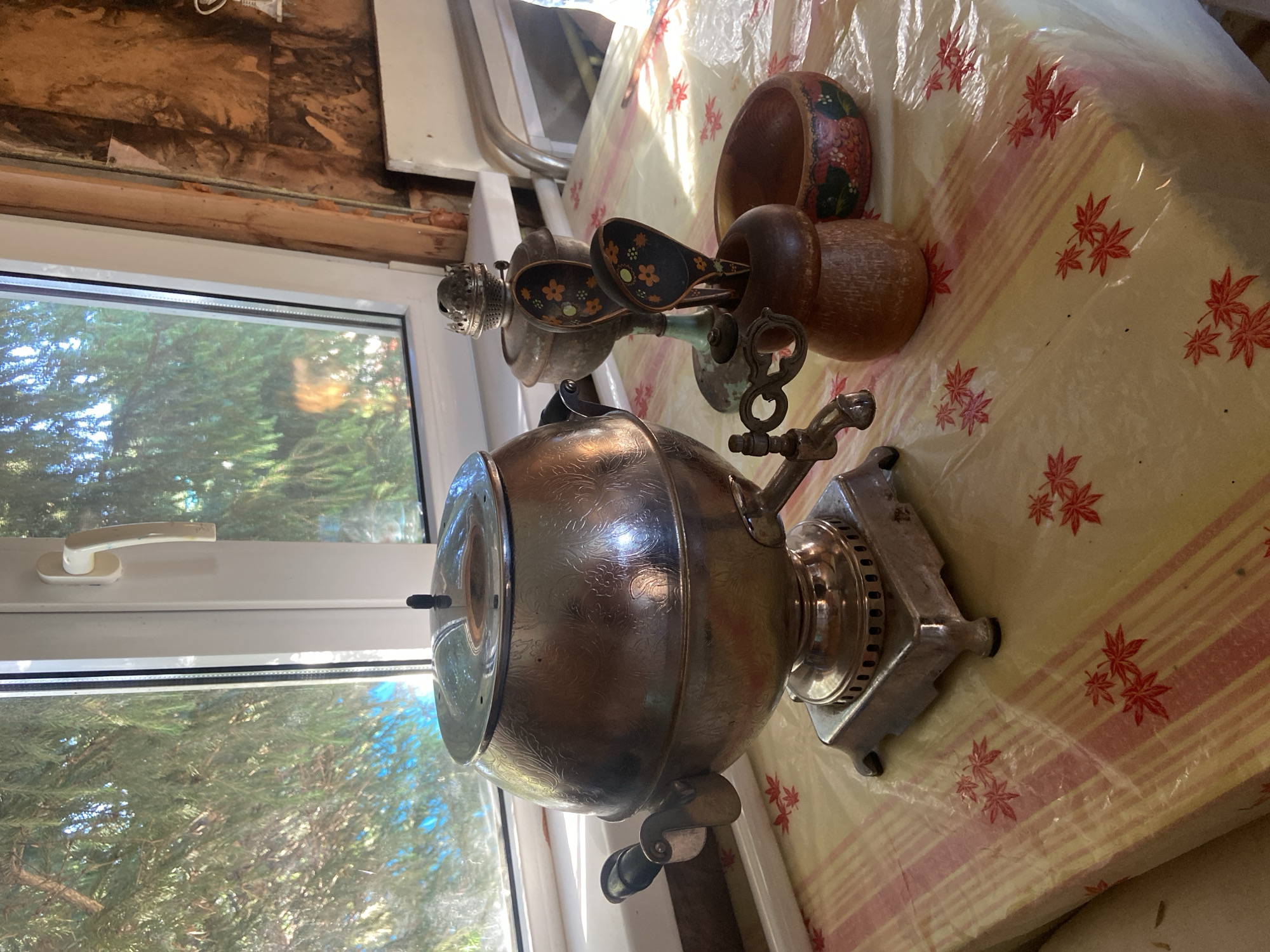 A close-up, eye-level shot showcases a domestic scene featuring traditional Russian tea-making items positioned on a tablecloth near a window. The primary subject is a large, ornate metal samovar, characterized by its spherical body, decorative engraving, spigot, and handles. To its right, a smaller metal oil lamp with a decorative top and a wooden holder containing two decorative wooden spoons are visible. A small wooden bowl with colorful floral painting is also present. The items rest on a yellow and pink striped tablecloth adorned with red leaf patterns. The background reveals a window with green foliage outside and a portion of a wall with exposed wood framing and insulation. The setting appears to be indoors, possibly a kitchen or living area, in Naberezhnye Chelny, Russia. There are no people or animals present in the image. The lighting suggests it is daytime, with sunlight streaming through the window, casting reflections on the metal objects.