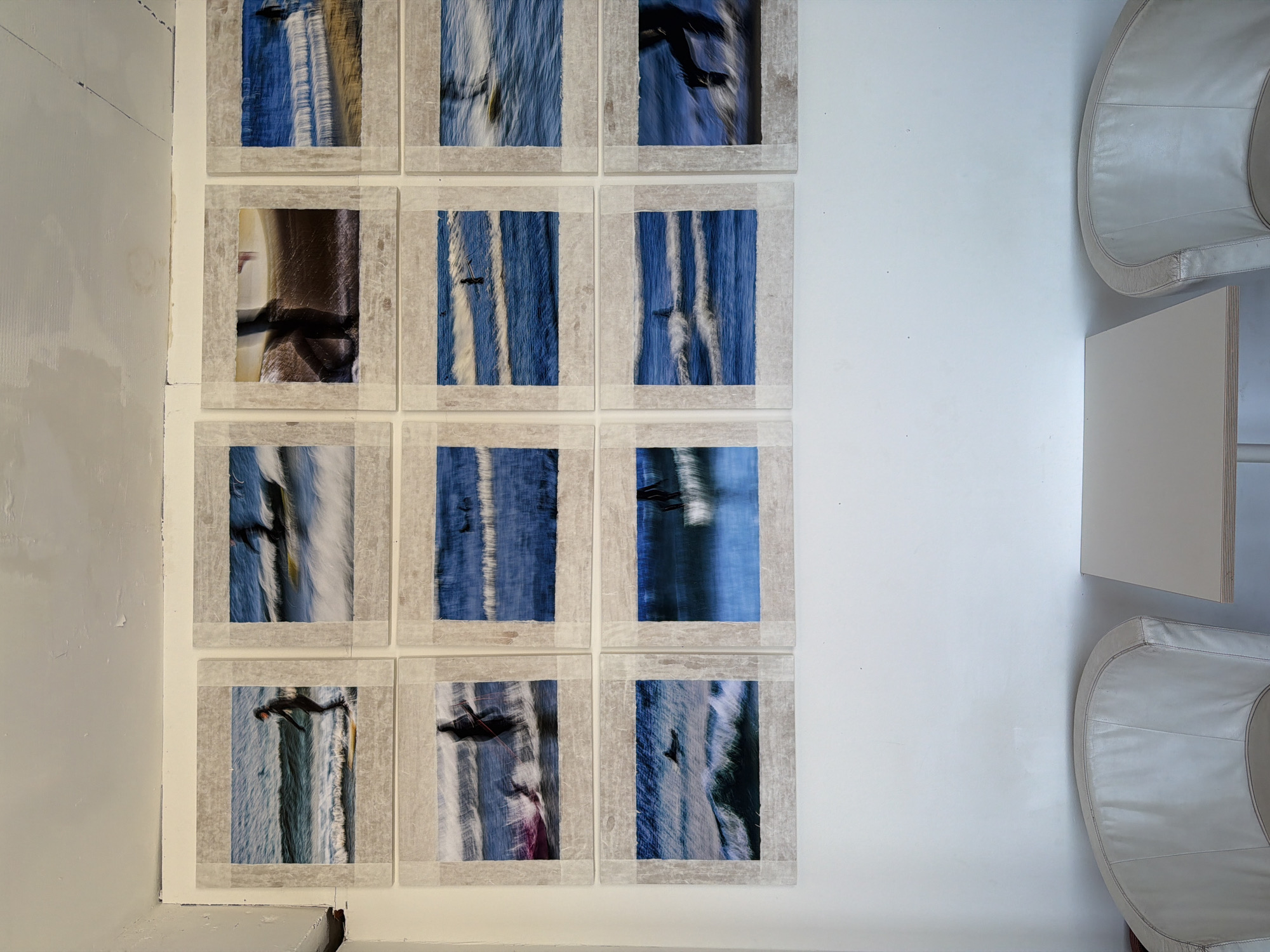 Twelve square photographic prints, framed in a pale gray border that appears to be secured with tape, are arranged in a grid of three rows of four on a white wall. The prints primarily contain images of ocean waves and surfers, with varying degrees of blur indicating movement. One print in the top-left corner features a close-up of a person.
To the right of the photographs, two rounded white chairs are positioned, flanking a low white rectangular table. The walls are painted white, and the left wall has evidence of repairs. The setting appears to be a gallery or living space in Hyeonnam, South Korea.