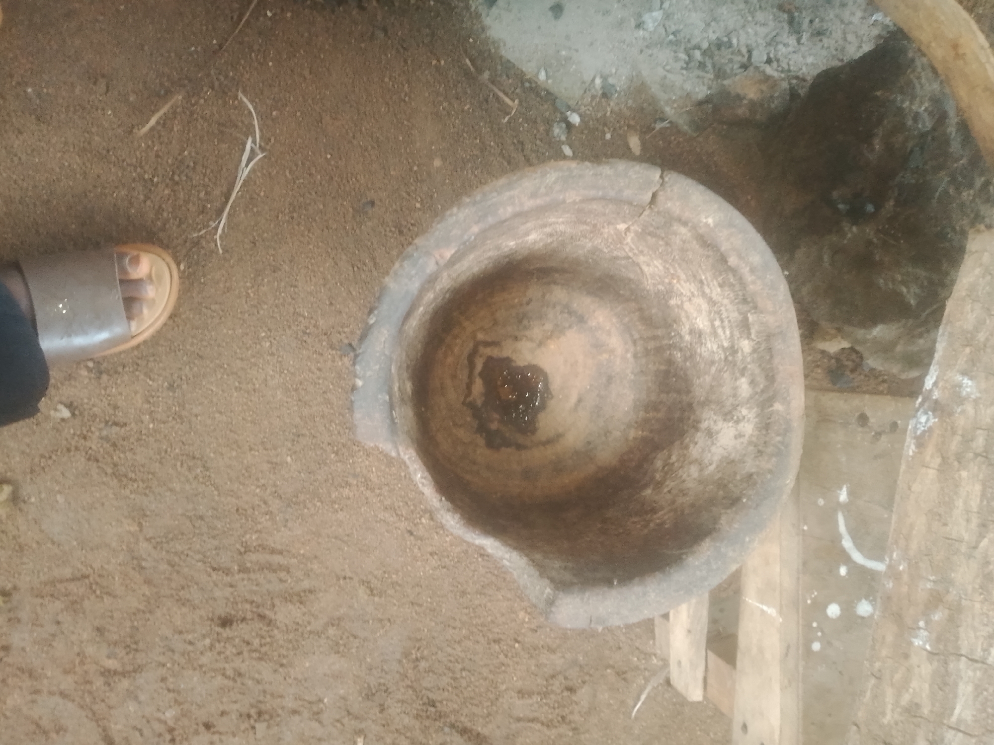 In Jalingo, Nigeria, a medium shot captures an overhead view of a traditional mortar with some dark liquid residue inside, resting on sandy ground. To the left, a person's foot wearing a brown sandal is visible. The background consists of more sandy ground and rough, natural materials including rocks and wood. The scene is illuminated by natural light.