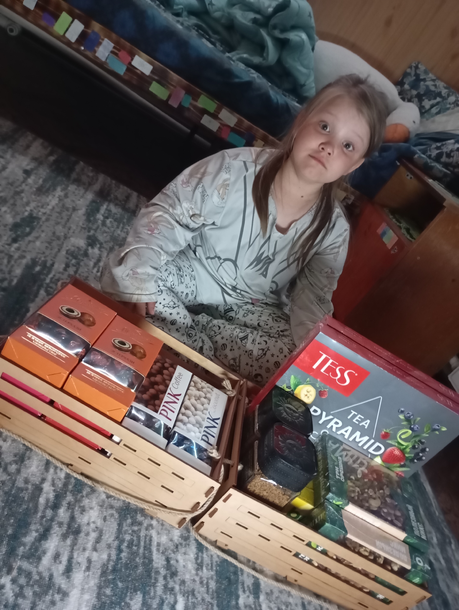 A young girl sits on a patterned rug with two wooden crates containing food and beverages in an indoor setting.
The girl, who appears to be about eight years old, is wearing patterned pajamas, a light-colored, long-sleeved shirt with dark lines, and has her light brown hair pulled back in a loose ponytail. She looks toward the viewer with a neutral expression.
In front of her are two wooden crates, filled with various items. The crate on the left contains brown and orange boxes with "KTEPXAЛИ" printed on them, boxes labeled "PINK Coffee," and "PINK Coconut." The crate on the right contains a red box labeled "TESS TEA PYRAMID," black containers, and other green and white boxes containing coffee.
Behind the girl is a bed with a blue and white comforter and some furniture with colorful decorations. The rug beneath them has a dark, patterned design.