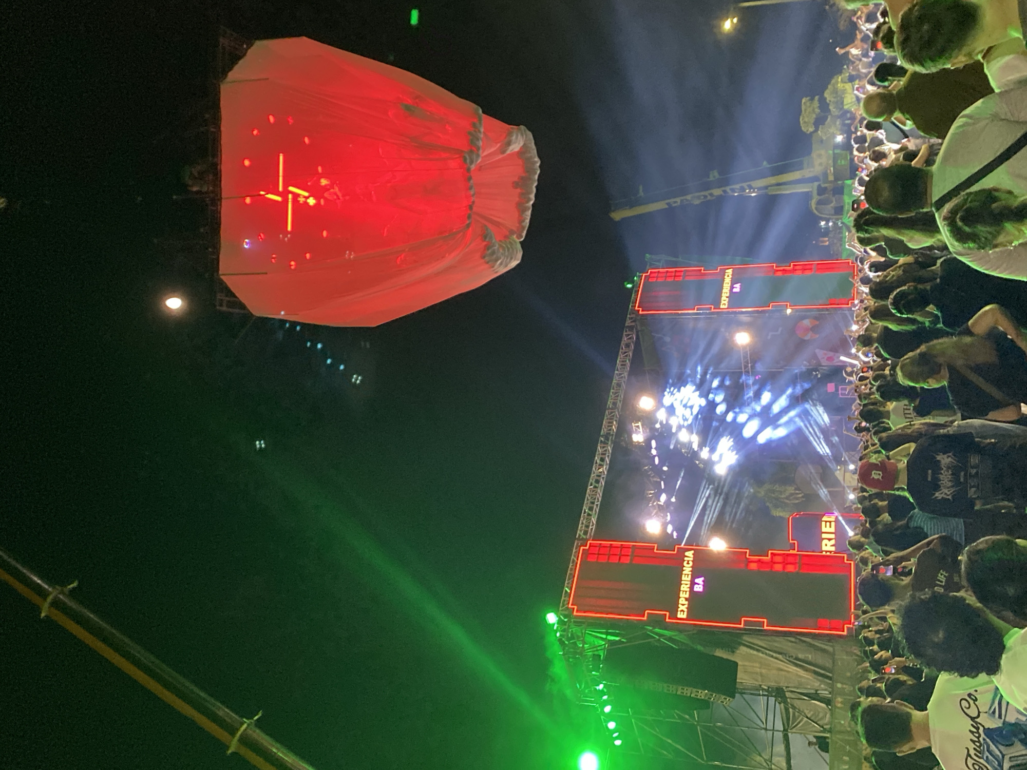 The image shows a large crowd gathered at an event in Buenos Aires, Argentina. A large red Chinese lantern floats above the audience. A stage with the words "Experiencia BA" is visible in the background. The environment is illuminated by stage lights and beams. The crowd is dense, with many people facing the stage and likely engaging with the event.