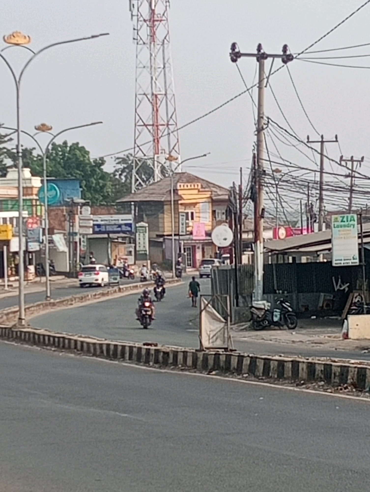 The image depicts a street scene in Bandar Lampung, Indonesia, likely on a cloudy day. A white car is parked on the side of the road, facing away from the camera. Several motorcycles are on the road, some with riders. A person in dark clothing is walking away from the camera on the sidewalk. Buildings with various signs and advertisements line the street. A tall communication tower is visible in the background, along with numerous electrical wires and poles. The road is paved and curves to the right. A concrete barrier with black and white stripes separates the road from the sidewalk area. There is a sign that reads "ZIA Laundry".