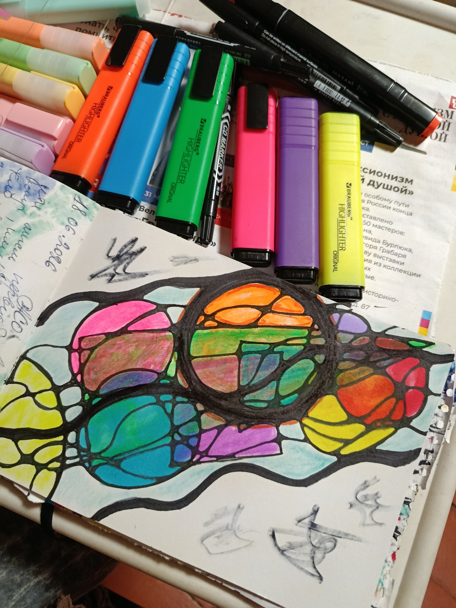The image displays a close-up view of a desk or workspace with art supplies and a partially completed drawing. Several brightly colored highlighter pens are arranged on top of some papers, including orange, blue, green, pink, purple, and yellow. Black marker pens are also visible.
The central focus is a notebook page with a drawing created using a neurographic art technique. The drawing features organic, interconnected shapes filled with vibrant colors like pink, orange, green, blue, yellow, and purple, outlined in thick black lines. The style is abstract and dynamic, suggesting a creative process.
To the left of the drawing, there is some handwritten text and what appears to be a date: "18.06.2016". The background consists of newspaper clippings and other printed materials, providing a textured and layered setting. The overall impression is one of an artist actively engaged in their creative work, surrounded by their tools. The lighting suggests it might be indoors, possibly during daylight hours, but there are no strong indicators of a specific time of day or weather. The location is not discernible from the image.