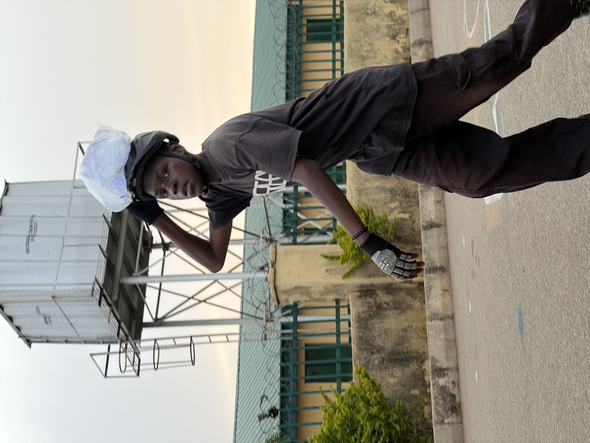 A young man wearing a helmet and gloves is standing on a paved surface next to a building with green barred windows. He is wearing a grey t-shirt and dark pants. A plastic bag is perched on top of his helmet. Behind him is a large metal water tank. The location is Jos, Nigeria.