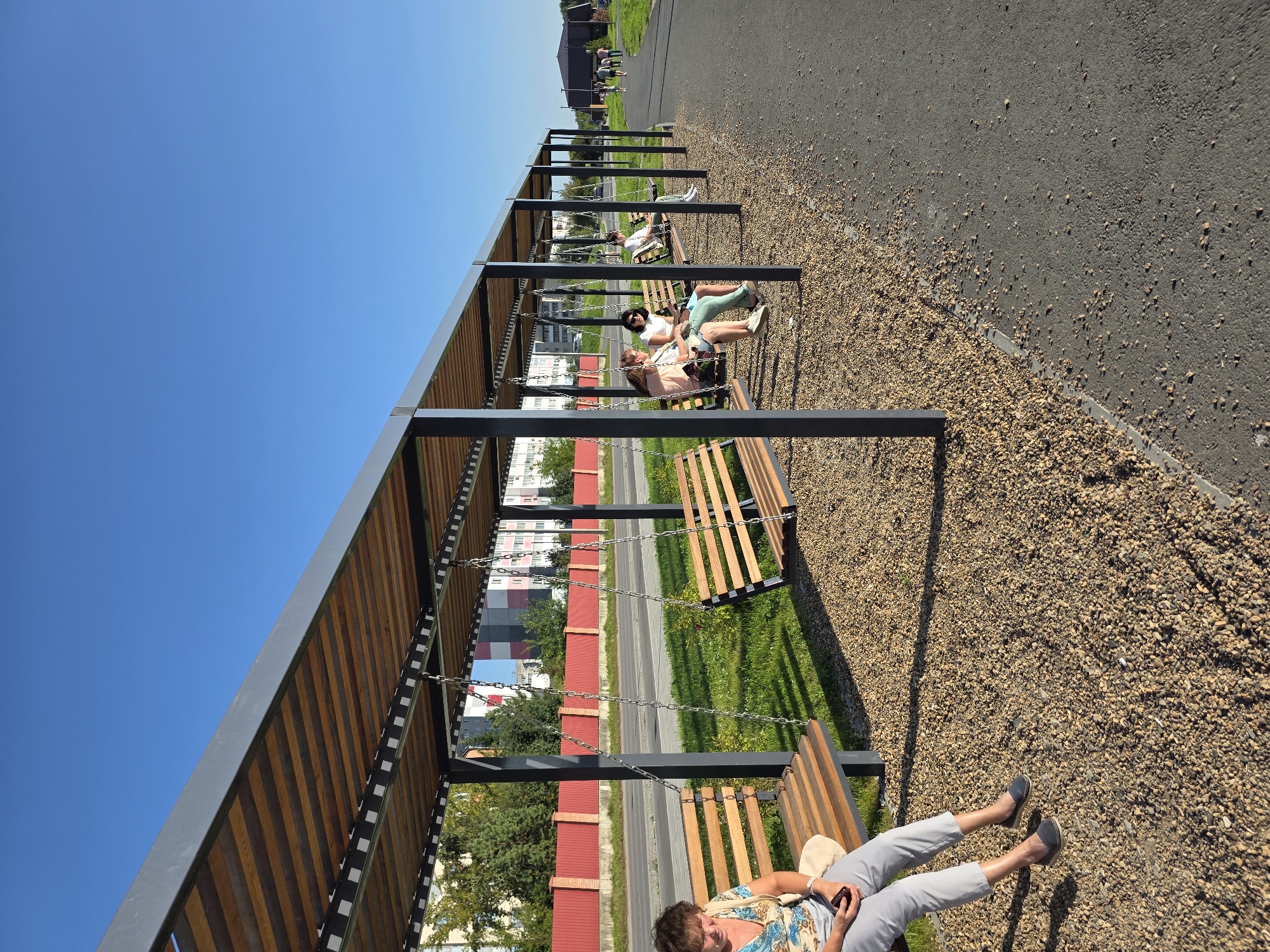 An outdoor scene in Miass, Russia, shows a long, covered walkway with a series of suspended wooden benches functioning as swings. The walkway is supported by dark grey metal posts and has a slatted wooden roof. The ground beneath the walkway is covered in small, light brown gravel, transitioning to a grassy area on one side and an asphalt pathway on the other.
In the foreground, a woman with dark hair, wearing a light-colored patterned top and grey pants, sits on the first swing bench on the left, holding a mobile phone in her right hand. She is looking down at the phone and appears relaxed.
Further down the walkway, other individuals are seated on the swings. One group of three young women is on a swing, with one holding a tablet. Another person is seated on a swing further back. In the distance, several people are walking along the asphalt path.
Behind the walkway, a bright red fence runs along the edge of a road. Beyond the fence, there are trees and multi-story buildings. The sky is clear and blue. The overall impression is one of a public recreational space on a sunny day.
