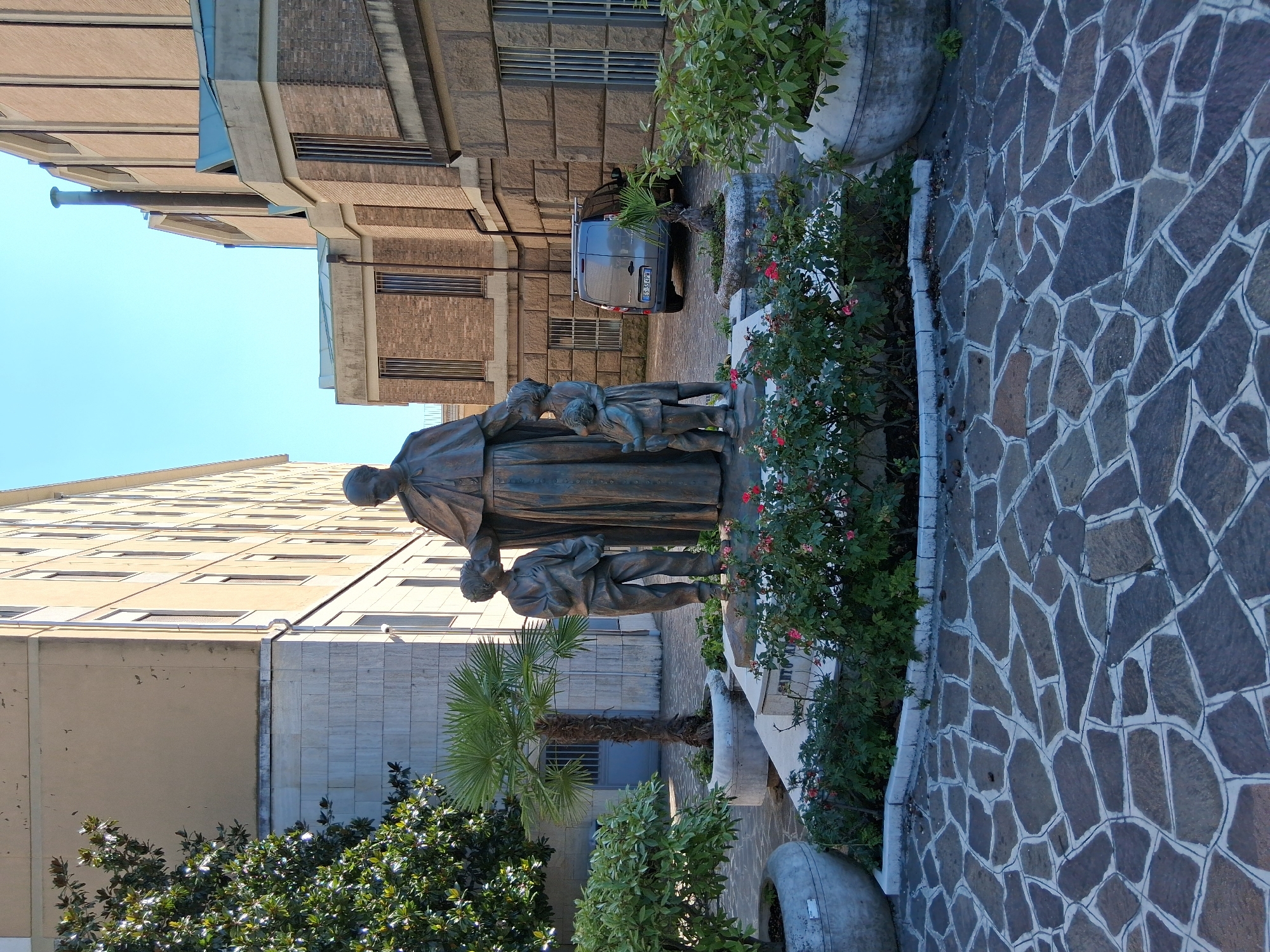 This is a full-shot, eye-level, outdoor, daytime photograph of a bronze statue of a robed male figure with two children, one on each side, in a courtyard setting. The statue is positioned in the foreground, centrally framed, on a raised platform surrounded by a flower bed. The male figure, possibly a priest or religious figure, stands tall with one hand resting on the head of the child to his right. The child on his left holds a book. The children appear to be looking up at the man.
The courtyard is paved with irregular, flat stones of various shades of gray and brown, set with white grout. To the left of the statue, a large, grey, circular planter contains a leafy green shrub. Further left, a palm tree in a similar planter stands tall. Behind the statue and flower bed, a beige, multi-story building with numerous windows occupies the left side of the frame. To the right of this building, a brick structure with a flat roof and metal-framed windows is visible. In the background, a dark grey van is parked. Further to the right, another brick building with a more modern architectural style featuring large windows and a blue roof section is present. The sky is clear and blue.
The location is identified as being in Padua, Italy, likely within the grounds of a religious or educational institution given the presence of religious iconography and the architectural style of the buildings. Specifically, this image depicts the exterior of the Collegio Gregorianum in Padua, Italy.