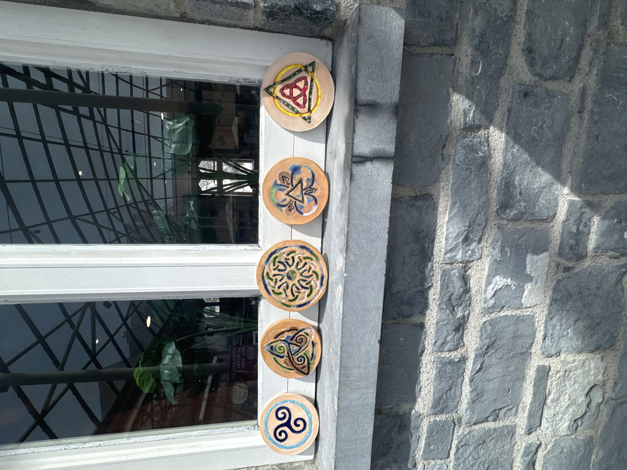 Five circular ceramic coasters with Celtic knot designs are displayed in a row on a stone windowsill. The coasters have distinct patterns in various colors, including blue, green, brown, yellow, and red. The background shows a window with a reflection of greenery and an interior space. The windowsill rests on a dark gray stone wall.