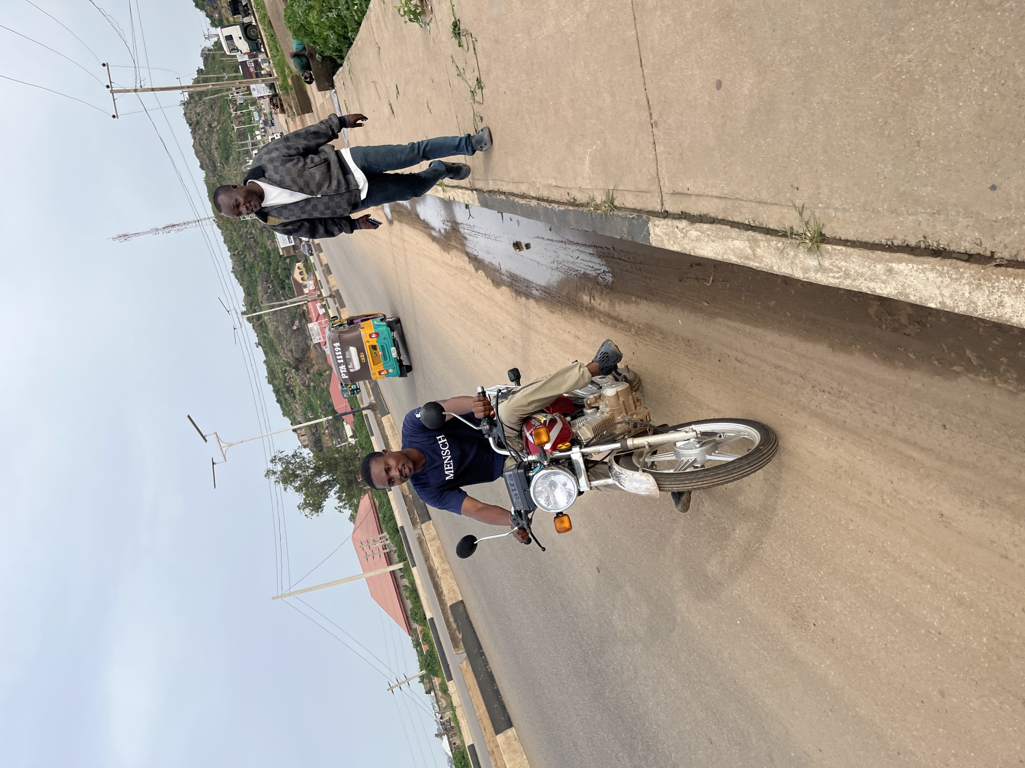 The image depicts a street scene in Gura Top, Nigeria. Two men are visible. One man, wearing a checkered jacket over a white shirt and blue jeans, stands on a concrete sidewalk, looking towards the camera and smiling. He holds a mobile phone in his left hand. The second man is seated on a motorcycle, wearing a dark blue t-shirt with white text and light-colored trousers. He is also looking at the camera. The motorcycle appears to be red and silver. In the background, a yellow and green motorized tricycle is visible. The setting includes a paved road with a curb and sidewalk, power lines, and hilly terrain with sparse vegetation and some buildings in the distance. The sky is overcast, suggesting a cloudy day. There is a wet patch on the sidewalk, possibly from recent rain or a spill. The visible text on the second man's t-shirt reads "MENSCH". The tricycle has text that appears to be a license plate number. The scene appears to be during daylight hours.