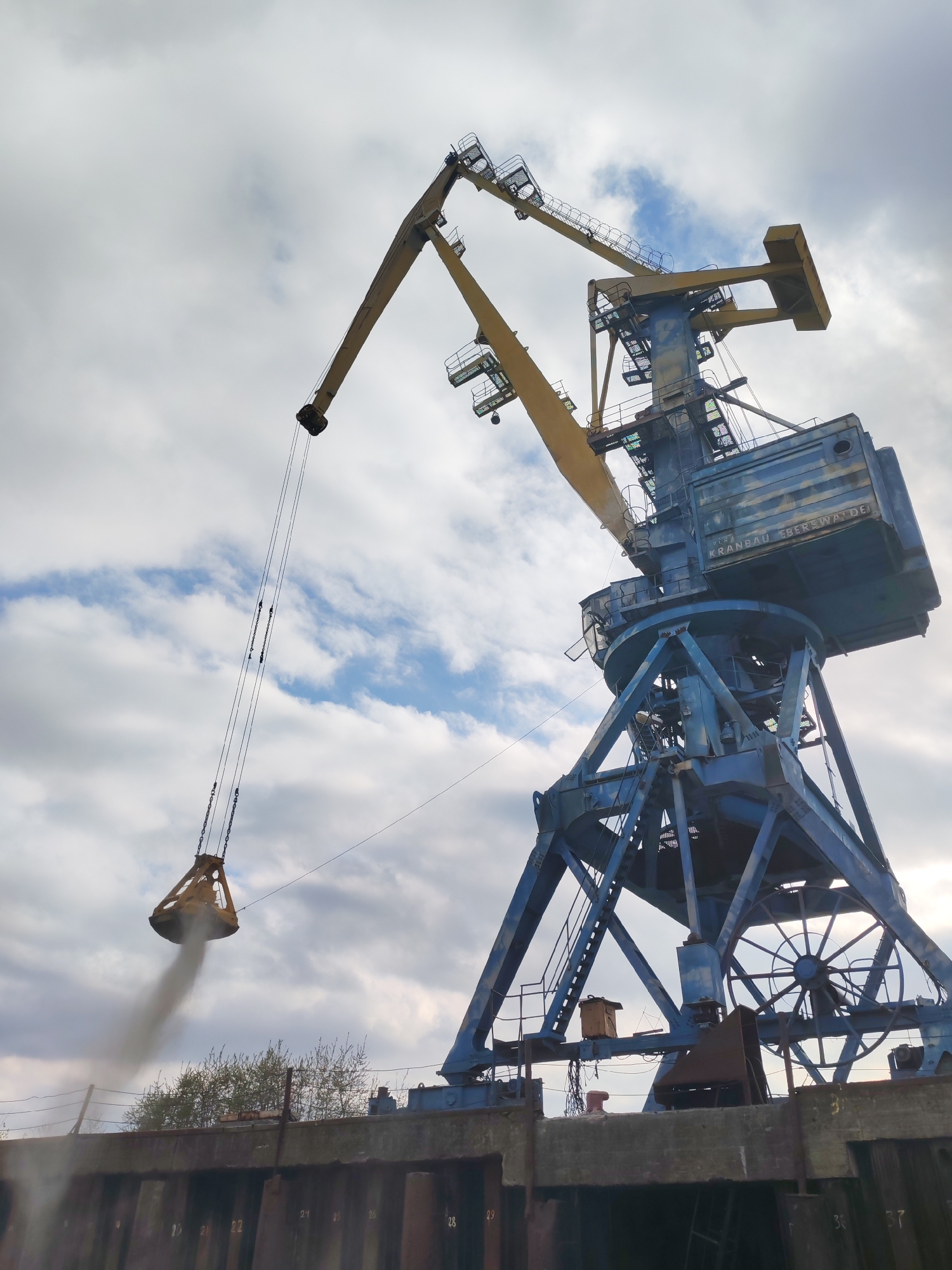 The image shows a large, blue and yellow harbor crane operating on a concrete dock. The crane has a clamshell bucket suspended from its boom, releasing dark material, likely sediment or debris, into the water or onto a surface obscured by the resulting cloud. The crane is labeled "VEB KRANBAU EBERSWALDE". The dock has metal pilings or sheet piling along its edge. Behind the dock is a line of small trees or shrubs. The sky is overcast with patches of blue.
While the crane's design suggests it's from the VEB Kranbau Eberswalde, a company from the former East Germany, I do not have specific information to confirm it is located in Tver, Russia. Additional context would be required to identify it with absolute certainty.