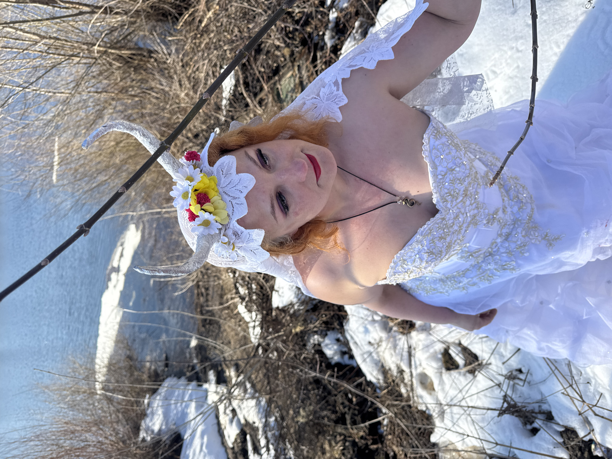 A woman in a white dress and headdress with horns poses outdoors in a snowy, wooded area. She has red hair, red lipstick, and wears a pendant necklace. Her dress is embellished with white and gold details, and the headdress features white lace and artificial flowers. The background includes a body of water and bare branches, indicating a wintery or early spring environment. The photograph's location is specified as Chaikovsky, Russia.