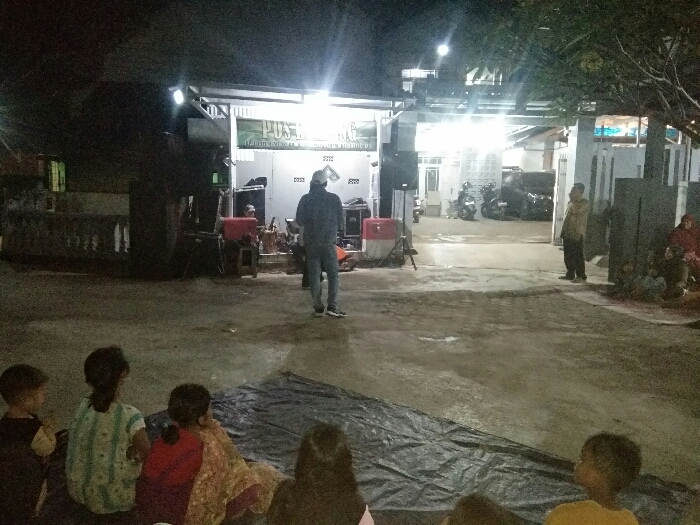 An outdoor night gathering is captured in Baleendah, Indonesia, with a group of people facing a brightly lit stage-like area in front of buildings. In the foreground, several children are seated on a dark tarp spread on the concrete ground, their backs to the viewer, appearing to watch an activity.
In the mid-ground, a man in a dark jacket and jeans stands facing the lit area, with sound equipment, including large speakers and a red cooler, positioned around him. Another man in a light-colored shirt stands further right, near the buildings. The background features several brightly illuminated buildings, possibly residential or community structures, with multiple artificial lights creating significant glare. Parked motorcycles are visible in front of these buildings, and some foliage frames the scene from the right.
The visible text on a banner or sign on the main building reads "PUSKESM", likely indicating a "Puskesmas" or community health center, suggesting the event might be related to public health or community welfare. The scene depicts an informal community event or gathering, with participants focused on the central illuminated area, possibly for a performance, presentation, or screening. The overall atmosphere suggests a communal night activity.