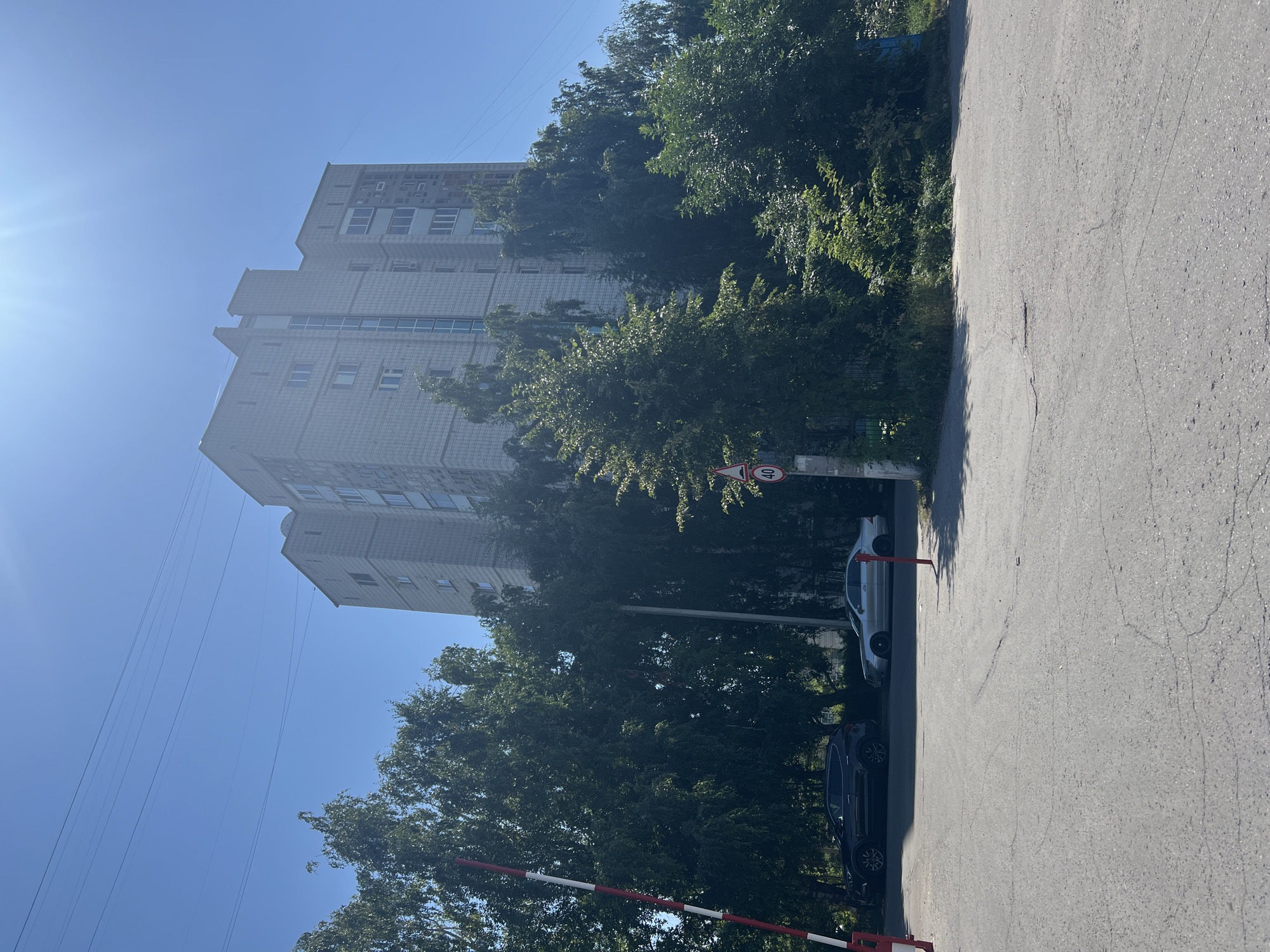 The image depicts a view of a large, multi-story apartment building in Ulyanovsk, Russia, situated behind a dense line of trees. The building is light gray with a textured facade and features numerous windows. A blue-tinted section is visible in the center of the building, possibly an elevator shaft or stairwell. The sky is clear and bright blue, with the sun strongly illuminating the scene from the upper left, creating lens flare.
In the foreground, there is an asphalt road with visible cracks and some wear. To the left, a red and white striped barrier gate is partially visible, suggesting a controlled access point. Two cars are parked along the edge of the road, partially obscured by trees. The car on the left is a dark SUV, and the car on the right is a silver sedan. A traffic sign with the number "40" inside a red circle, indicating a speed limit, is mounted on a pole to the right of the road. The overall environment appears to be a residential area with significant greenery surrounding the buildings. There are no people visible in the image.
