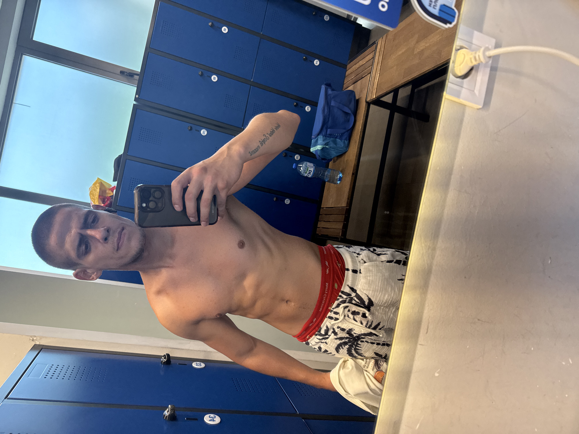 In a locker room, a shirtless man with a buzz cut takes a mirror selfie. He wears patterned shorts and red boxer briefs. To his right, a blue gym bag and a water bottle rest on a wooden bench. Behind him, a row of blue lockers lines the wall. The scene is illuminated by natural light streaming through a window above the lockers.