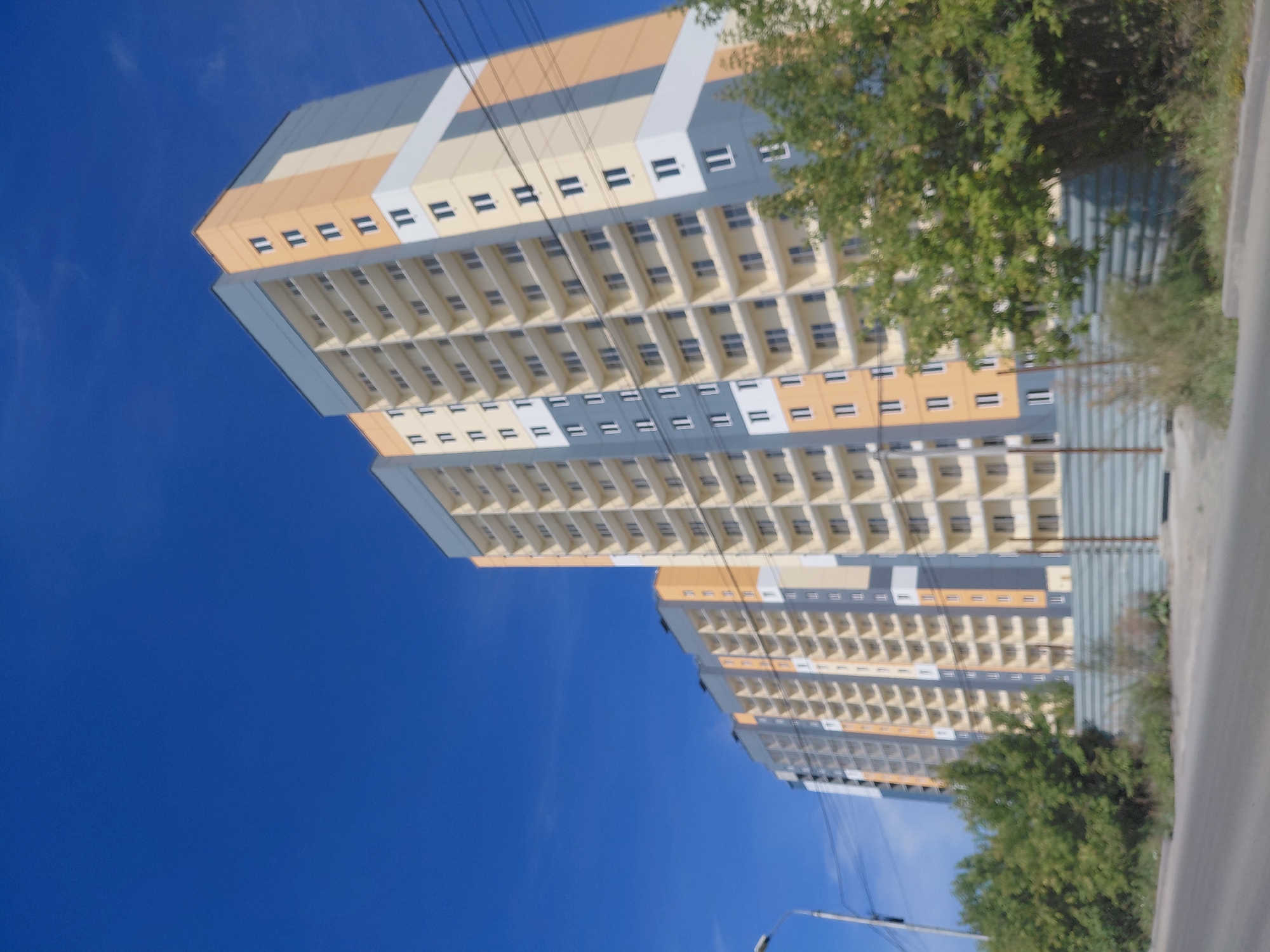 This is a vertical, eye-level, slightly tilted shot of a modern apartment complex under a clear, bright blue sky in Novosibirsk, Russia. The building, which appears to be under construction or recently completed, has a multi-colored facade in shades of pale yellow, grey, and light orange. It features numerous balconies with light-colored railings and uniformly placed windows. The building is large, with multiple stories, and appears to be a high-rise. In the foreground, there is a grey metal fence, some green foliage, and a glimpse of an asphalt road. Power lines are visible against the sky. The overall impression is one of urban development in a sunny environment.