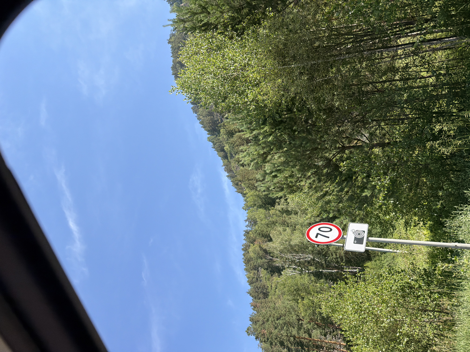 The image shows a road sign indicating a speed limit of 70, accompanied by a camera symbol, likely denoting a speed camera. This sign is situated in a natural environment, with a dense forest covering a hillside under a clear blue sky with sparse clouds. The foreground features lush green vegetation, including tall trees and bushes. The scene appears to be on the outskirts of a settlement, possibly in a rural or mountainous region. The presence of the speed limit and camera suggests a road is nearby, likely passing through this scenic area. The image could be captured from a vehicle, given the framing from what appears to be a window. The location is identified as Ust'-Muny, Russia.