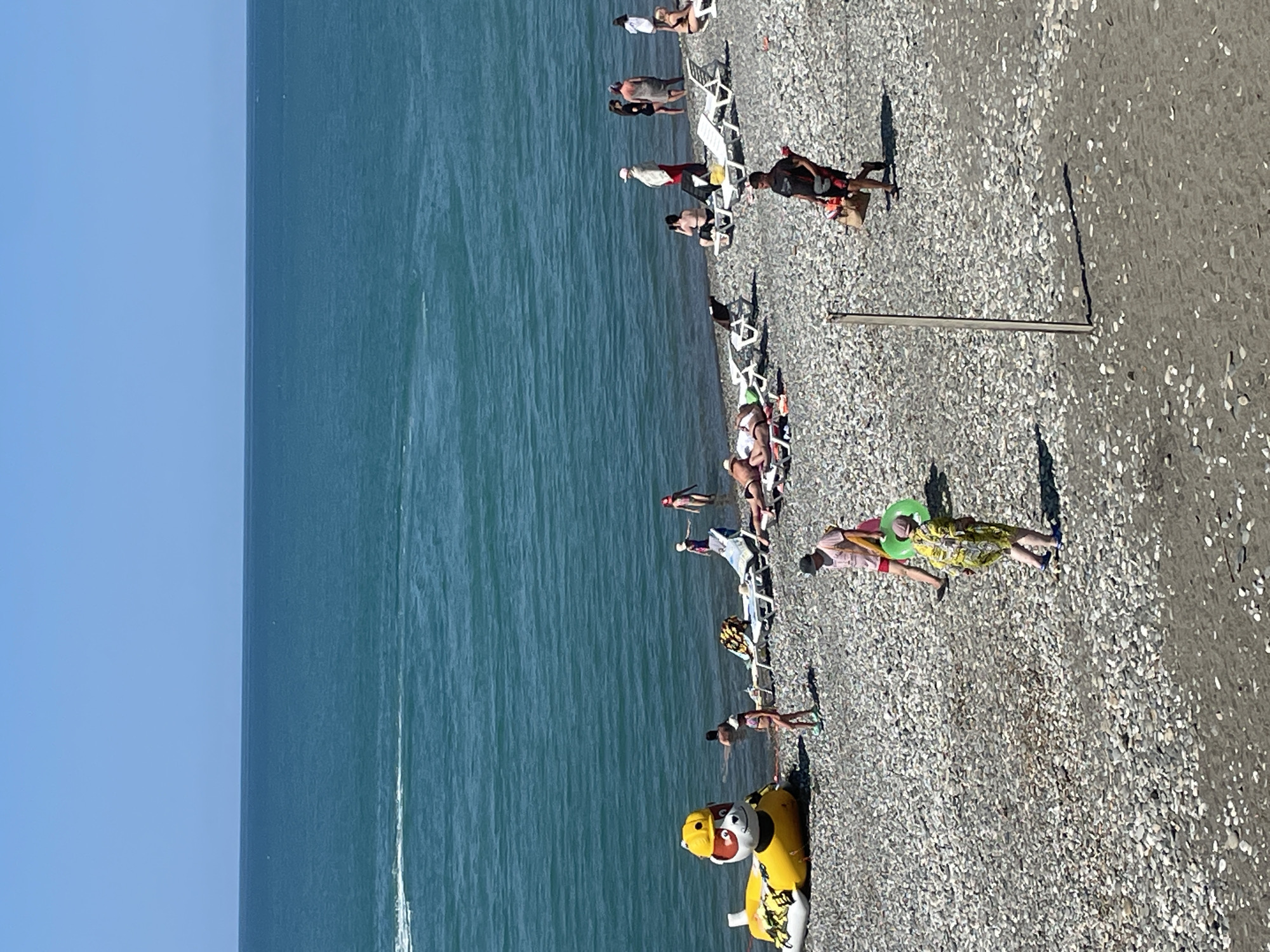 The image shows a crowded beach in Giachrypsh, Abkhazia. The sea is a bright turquoise color and appears calm. The beach is covered in grey pebbles and there are several people enjoying the sun and sea. In the foreground, a person in a yellow and green patterned outfit walks with a green inflatable ring. Behind them, a person in a pink t-shirt and red shorts walks with a bag.
Further back, there are people sunbathing on lounge chairs and wading in the water. To the left, a large inflatable yellow duck-like toy is partially submerged in the water. There are also several beach umbrellas and chairs scattered across the pebbles. In the background, the sky is a clear blue. The scene depicts a typical summer day at the beach with various recreational activities taking place.
