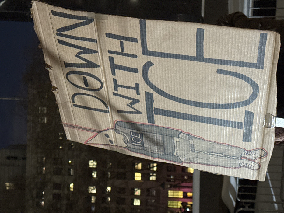 A cardboard sign reads "Down With ICE" in large, blocky, blue-gray letters. Beneath the text, a crudely drawn figure of a person is depicted lying horizontally. The figure is dressed in clothes labeled with "ICE." The sign has a tattered edge and is held up against an urban backdrop with buildings and lit windows visible in the background. The sign is slightly rotated, making the text appear vertically oriented.