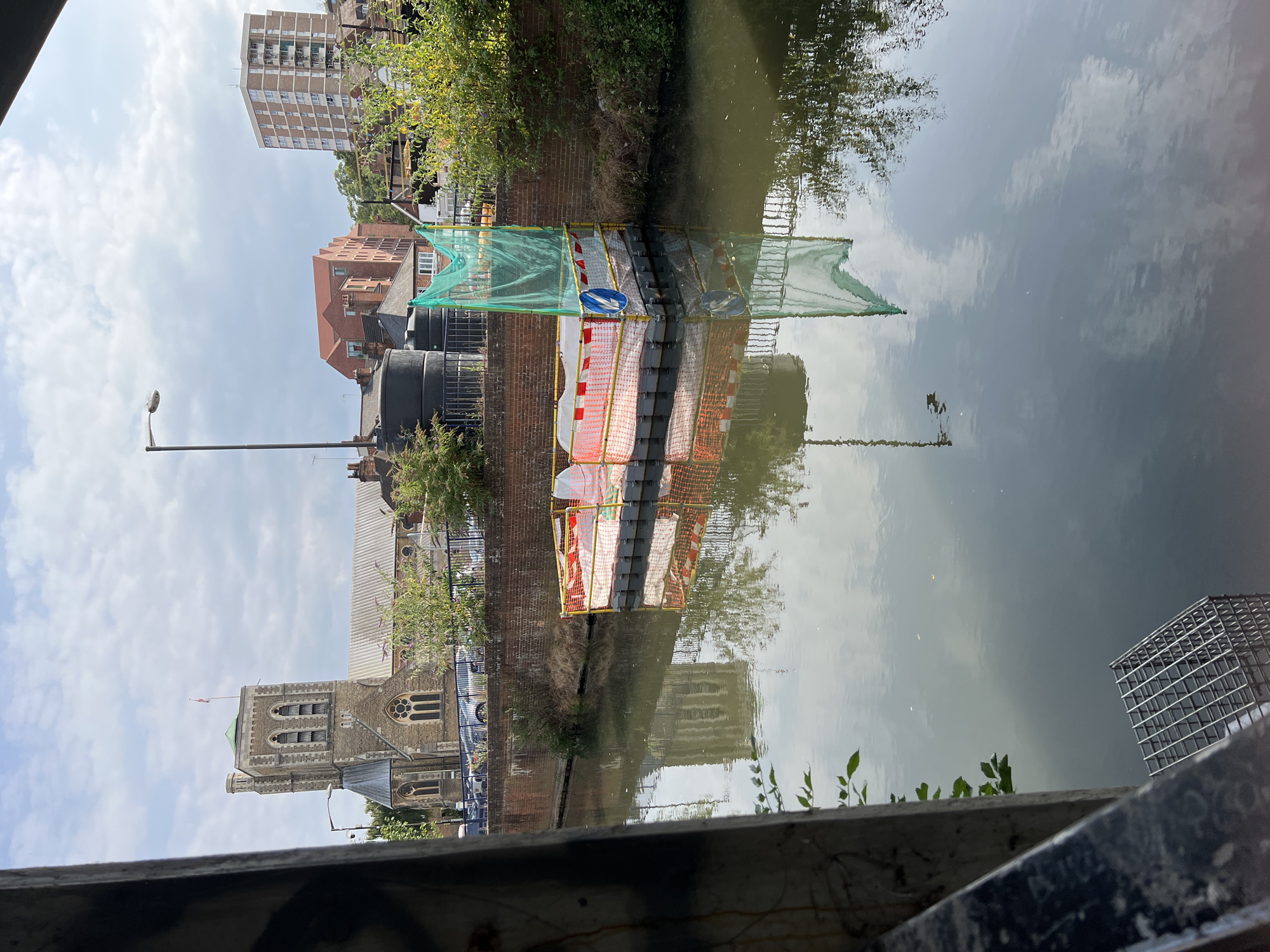 The image, captured on a cloudy day, depicts the River Wey in Guildford, United Kingdom, looking north. In the mid-ground, a temporary floating platform, likely for construction or maintenance, is moored in the river. It is surrounded by orange and white safety barriers and draped with a green net. A circular blue and white sign is attached to the platform.
To the left of the river, a brick wall with a metal fence runs along the embankment, behind which a church with a prominent clock tower and a red-tiled roof is visible. Several parked cars are discernible along the roadside adjacent to the church. Further in the background, a multi-story brick building with a red-tiled roof and a modern high-rise apartment building are visible. Lush green trees and foliage line the riverbank and the background. The water of the river reflects the sky, clouds, and the surrounding architecture. In the foreground, a dark, textured metal railing and a wire cage structure are partially visible, suggesting the vantage point is from a bridge or walkway.