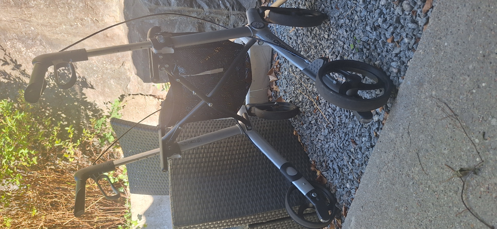 The image shows a gray rollator walker with a black mesh basket, parked on a gravel surface next to a concrete pathway. The walker has four wheels, two large ones in the front and two smaller ones in the back, and adjustable handlebars with grips. The background consists of a dark woven outdoor chair and lush green foliage, with a large rock formation to the right. The scene appears to be an outdoor setting, possibly a patio or garden area. The lighting suggests it is daytime, with sunlight casting shadows. There are no people or animals visible in the image. The specific location in Lillesand, Norway, cannot be determined from the visual information provided.
