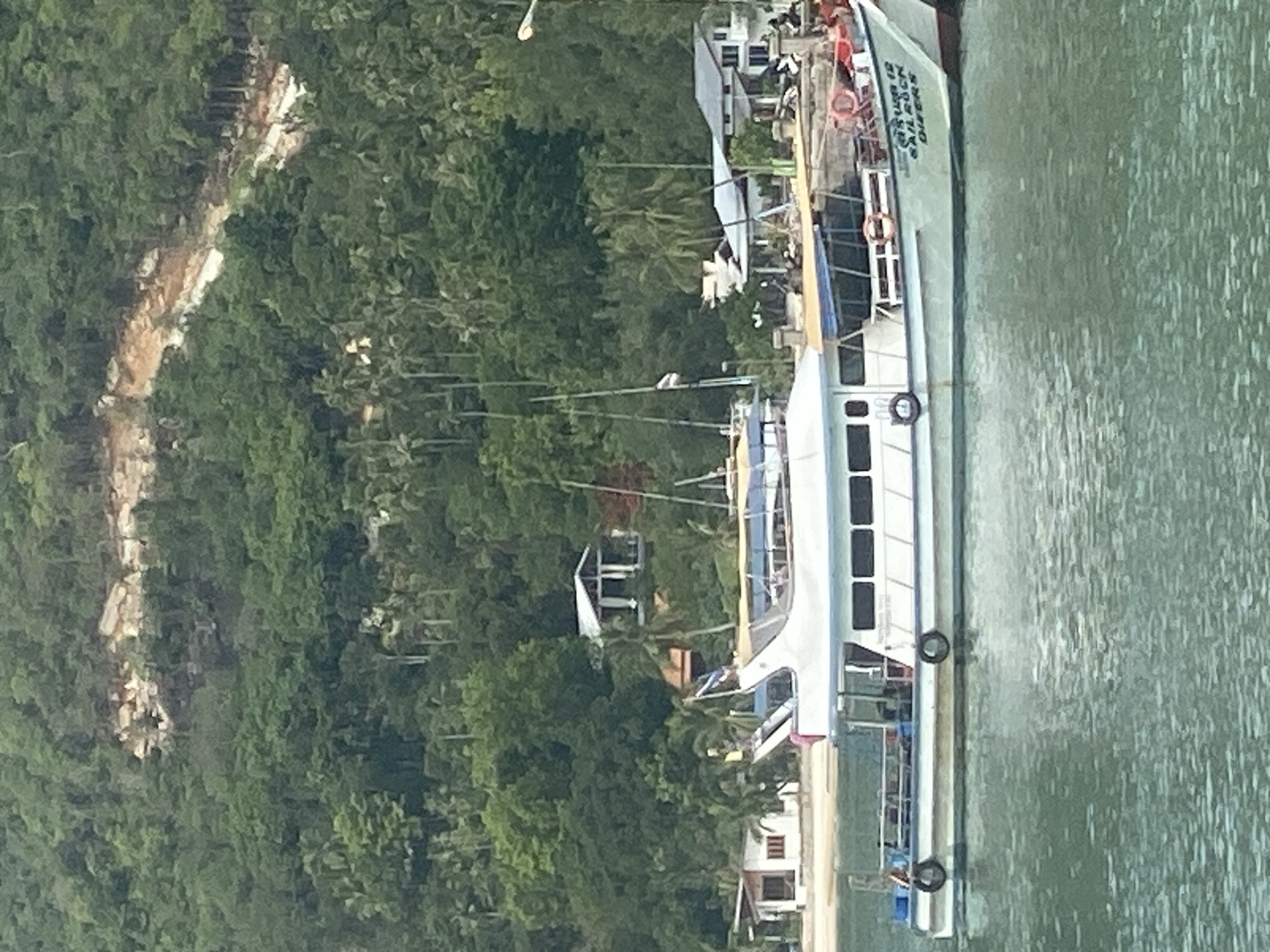 A landscape-oriented, daytime photograph captures a scene in Baan Chaloklum, Thailand. A multi-story boat is situated in the water along the shoreline. The boat's name "Sail Rock Divers" is visible. The boat is docked next to buildings with slanted roofing. Lush, green, dense forest covers the left side of the photograph. The water appears murky.