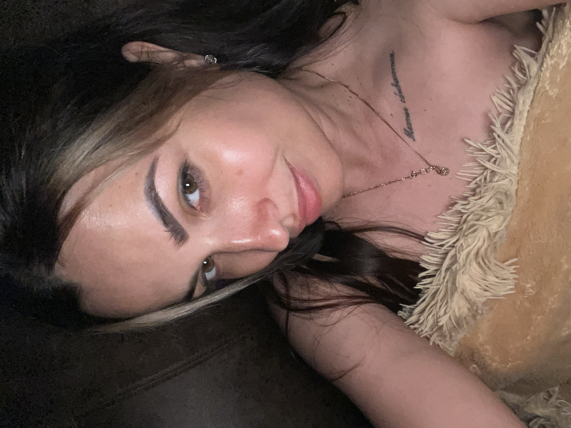 This is a vertical, close-up selfie of a young woman lying down, with her head tilted towards the camera. She has long, dark hair with some lighter streaks framing her face. She is wearing makeup, including eyeshadow and lipstick. A delicate gold necklace with a small charm is visible around her neck, and she has a tattoo on her upper chest that appears to read "Mami, cuéntame". She is wearing a ring on her left ear. The woman is lying against a dark surface, possibly a couch or bed, and is partially covered by a fluffy, light-colored throw blanket with a fringe. The lighting suggests an indoor setting, possibly in the evening or at night. Her expression is a relaxed, slightly playful smile.