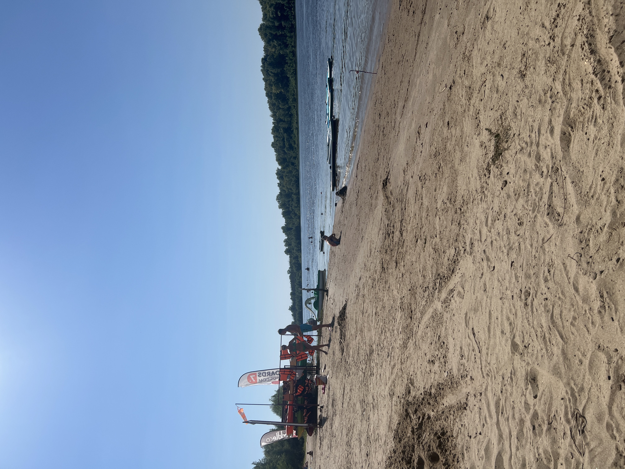 The image depicts a sandy beach along a wide, calm river under a clear blue sky. On the left side of the image, there is a rack with several kayaks and paddleboards, along with orange life vests. Two adults, a male and a female, are standing near this rack, appearing to be preparing for water activities. In the background, several paddleboards are floating on the river. Further back, a dense line of trees forms the opposite bank of the river. A few other people are visible scattered along the beach and in the water. The sand is a light brown color and shows some footprints and tire tracks. The overall scene suggests a recreational area during a sunny day.