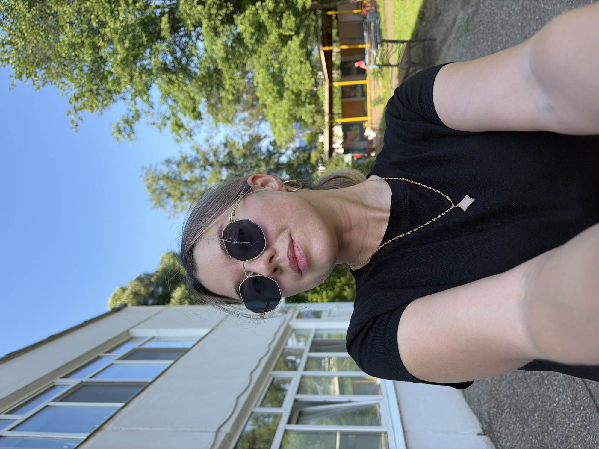 A young woman with blonde hair pulled back in a ponytail, wearing sunglasses, a black t-shirt, and a gold necklace with a diamond-shaped pendant, poses for a selfie outdoors. She is standing in front of a white building with multiple windows. In the background, there are trees, a blue sky, and a covered outdoor seating area with yellow pillars and benches. The image appears to have been taken in Chaikovsky, Russia, based on the architecture of the building and the style of the outdoor amenities, which are consistent with public spaces in the city. The lighting suggests it is a sunny day.