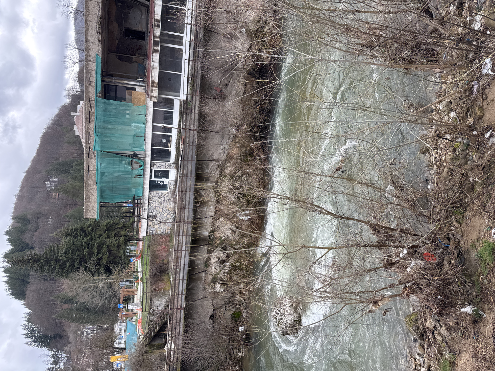 The image shows a view of a river flowing through Rudozem, Bulgaria. The riverbank is littered with trash. Next to the river is a multi-story building with windows and a stone facade, part of which is covered in green mesh. A metal railing runs along the edge of the river. The foreground includes bare branches and vegetation. In the background, a forested hillside is visible under a cloudy sky.