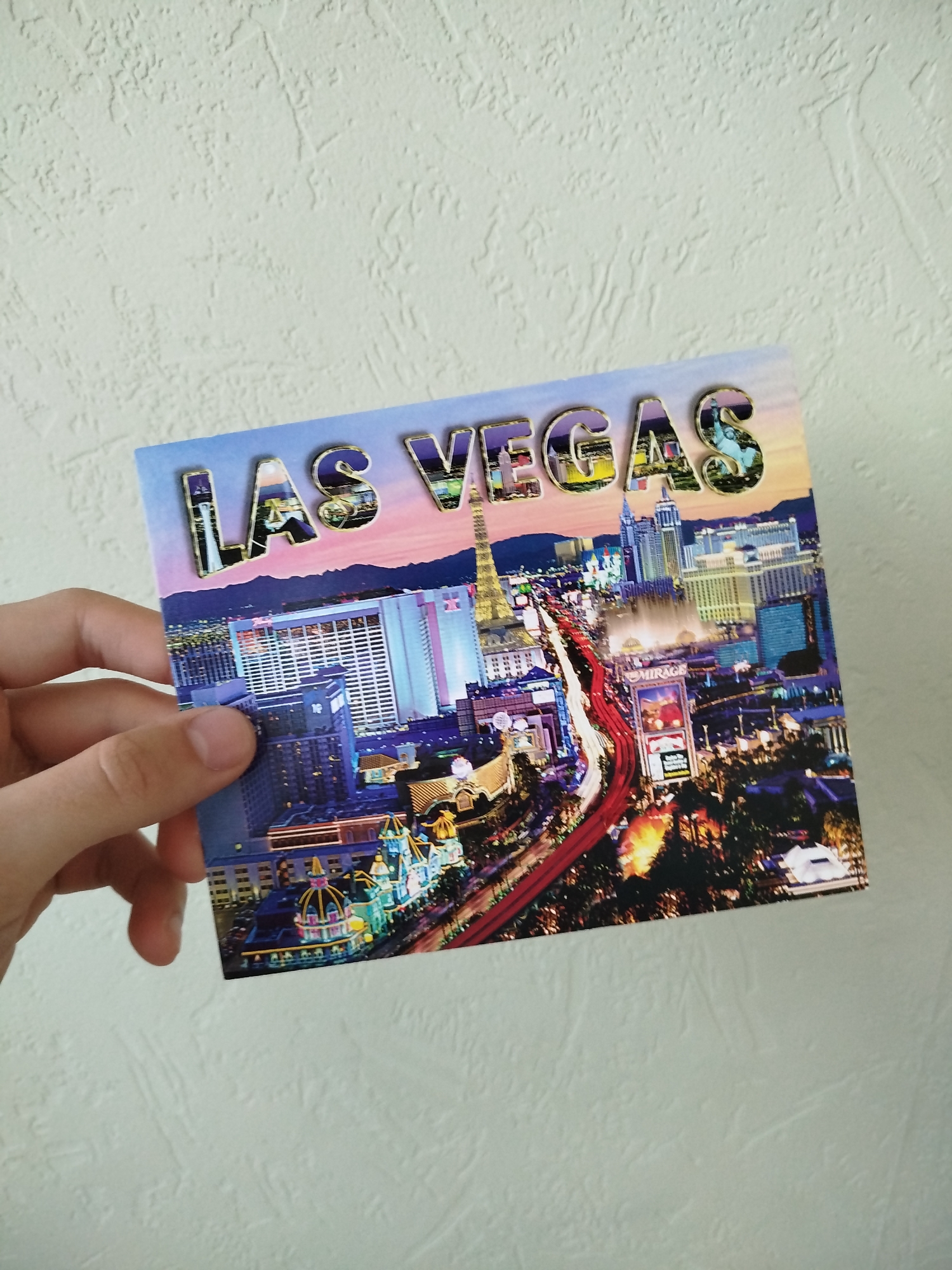 A hand holds a postcard of Las Vegas against a textured, light-colored wall. The postcard depicts a nighttime cityscape, with a highway leading into the city. The words "LAS VEGAS" are printed above the skyline, with the letters filled with corresponding city images.