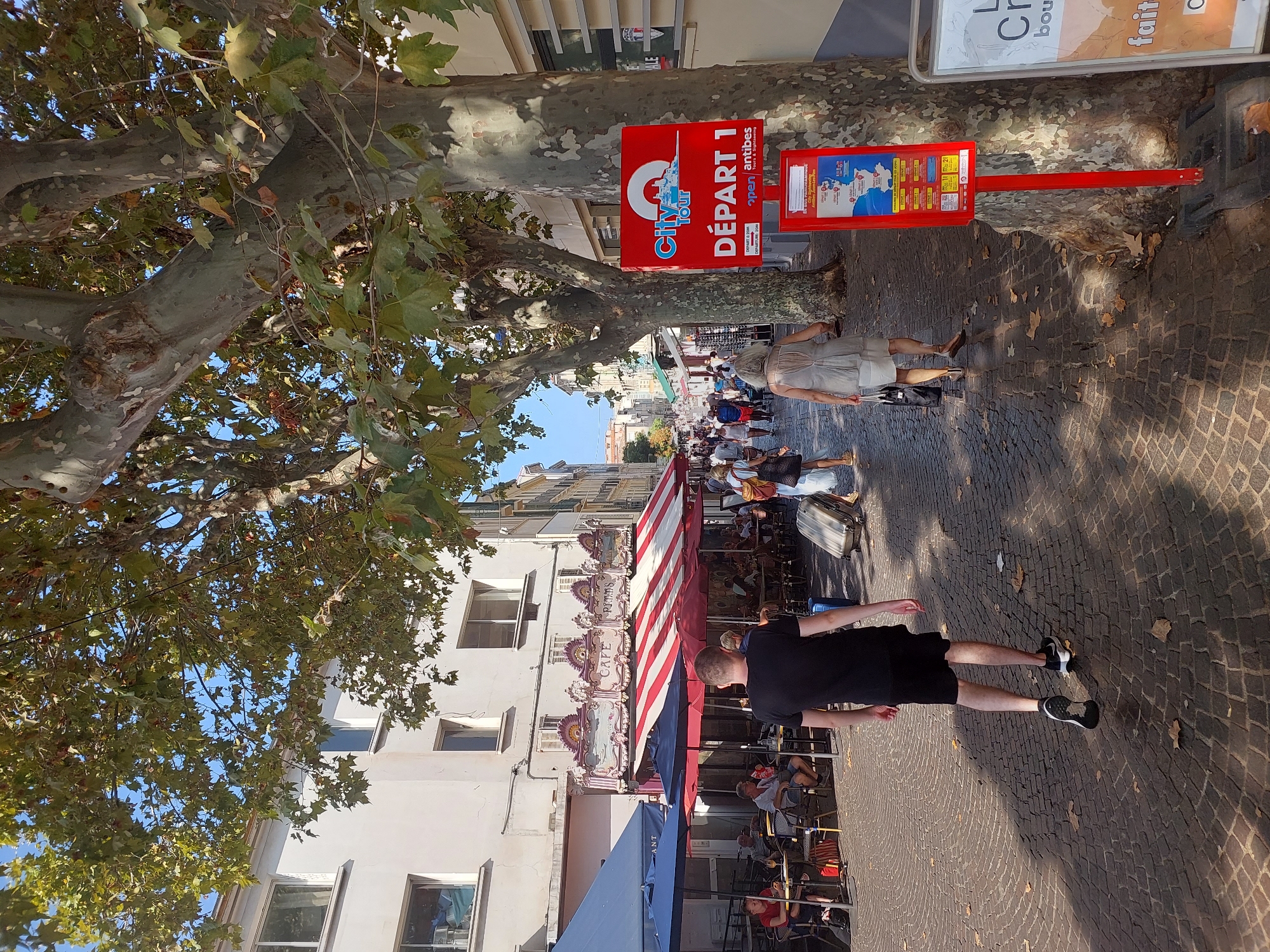 A street scene in Antibes, France, features a tree-lined cobblestone pedestrian walkway. On the left, buildings with open windows and awnings of blue and red and white striped fabric are visible, with tables and chairs set up outside a cafe. A carousel is partially visible in the background. On the right, a red signpost displays "City Tour DÉPART 1" and information about bus tours. Several people are walking along the street, some pulling luggage, and others seated at cafe tables. The sky is blue with some scattered clouds.