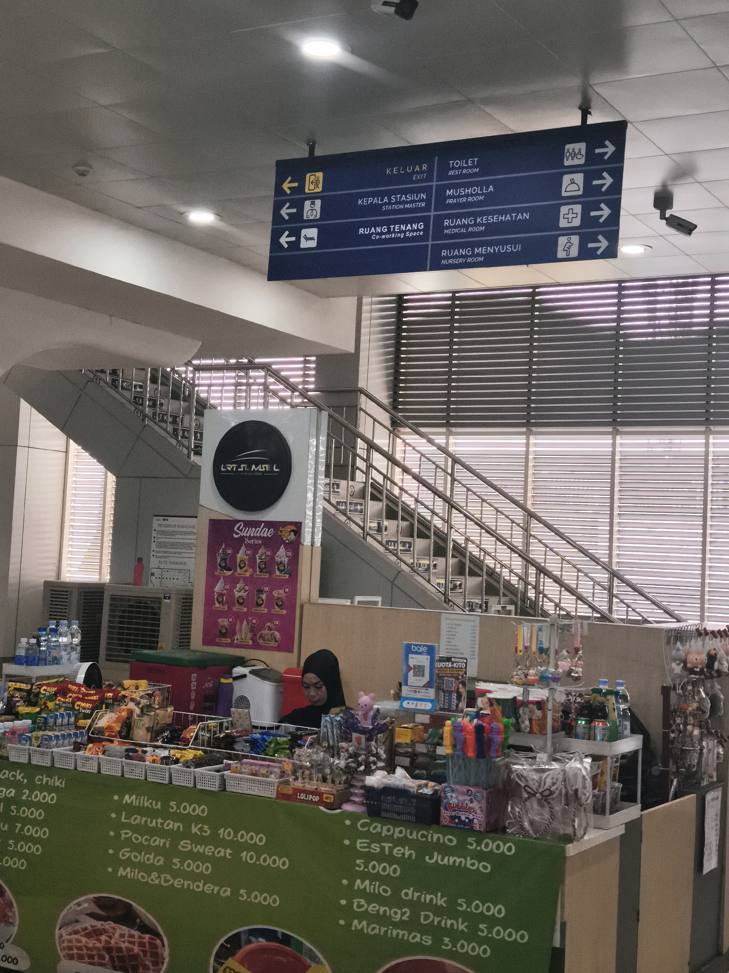 The image captures an interior scene, likely a public transit station, given the presence of directional signage and a small retail stall. A large blue directional sign is mounted on the ceiling, displaying directions in Indonesian and English. Destinations include "KELUAR (EXIT)", "TOILET (REST ROOM)", "KEPALA STASIUN (STATION MASTER)", "MUSHOLLA (PRAYER ROOM)", "RUANG TENANG (Co-working Space)", "RUANG KESEHATAN (MEDICAL ROOM)", and "RUANG MENYUSUI (NURSERY ROOM)", each with corresponding icons and arrows pointing to the right.
Below the signage, a staircase with metal railings ascends. To the left of the stairs, an air conditioning unit is visible. A circular black sign with white lettering, displaying "LRJ. MST L", is mounted on the wall near the staircase. A poster advertising "Sundae series" with various ice cream options is displayed prominently on a stand.
In the foreground, a retail stall is set up, selling various snacks, drinks, and small trinkets. A woman wearing a black hijab is seated behind the stall, facing towards the products. Bottles of water and various packaged snacks are neatly arranged on display shelves and in baskets. Some products have pricing listed on a green banner, including "Milk u 5.000", "Cappucino 5.000", and "Milo drink 5.000". A QR code is visible on a blue stand. The stall is illuminated by overhead lighting. The overall environment appears to be clean and organized, with a focus on public convenience and retail offerings. The location is specified as Kelurahan 16 Ilir, Kec. Ilir Timur I, Indonesia.
