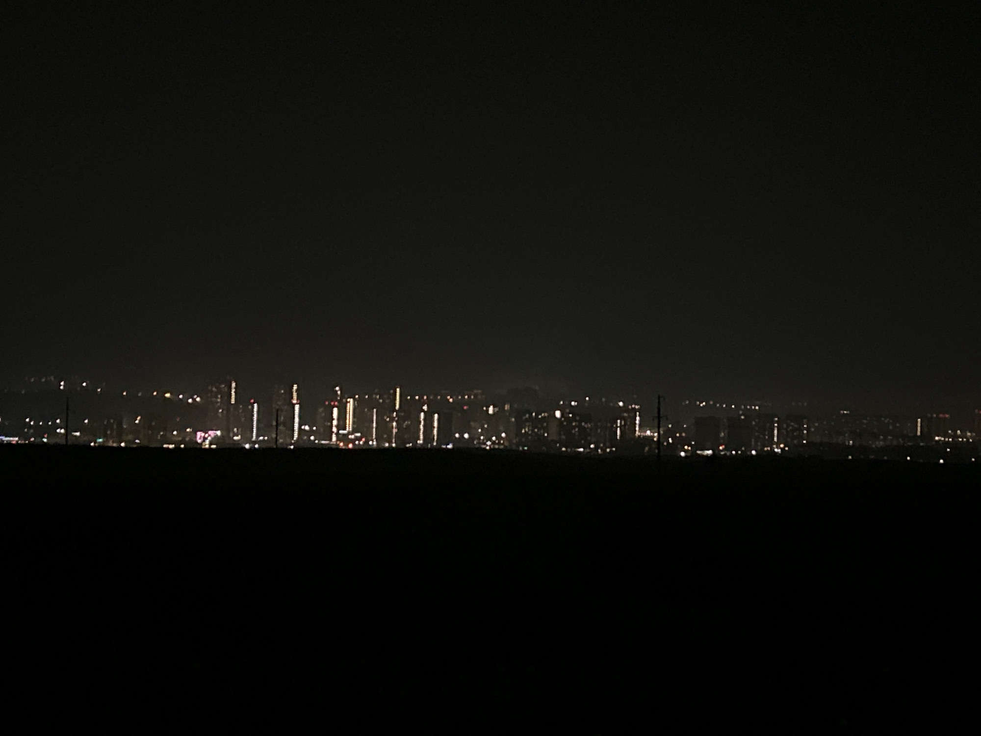 The image captures a distant, blurry view of a city skyline at night. The foreground is dominated by a large expanse of deep black, likely representing a dark field or unlit area. Above this, a faint line of lights stretches across the horizon, indicating the presence of buildings and infrastructure. These lights vary in brightness and color, with some appearing as small points and others as vertical streaks, suggesting tall structures like skyscrapers or illuminated windows. The sky is a dark, uniform color, with no visible celestial bodies or distinct cloud formations. The overall impression is of a city viewed from a considerable distance in low-light conditions, with the specific location in Solontsy, Russia, implied by the prompt.