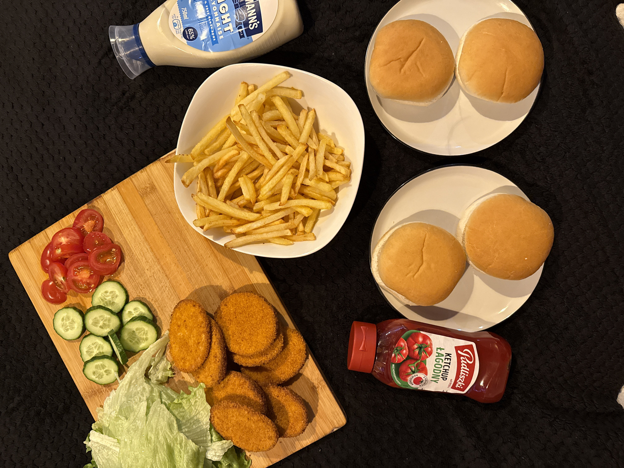 The image presents a flat lay arrangement of food items for making or consuming burgers. There are four burger buns arranged in pairs on two white plates. A white bowl holds a generous portion of French fries. A wooden cutting board features sliced tomatoes, sliced cucumbers, lettuce, and a stack of breaded patties. Condiments include a bottle of light mayonnaise and a bottle of ketchup. The entire arrangement is on a black textured surface. There is no indication the photo was taken in Great Yarmouth, United Kingdom.