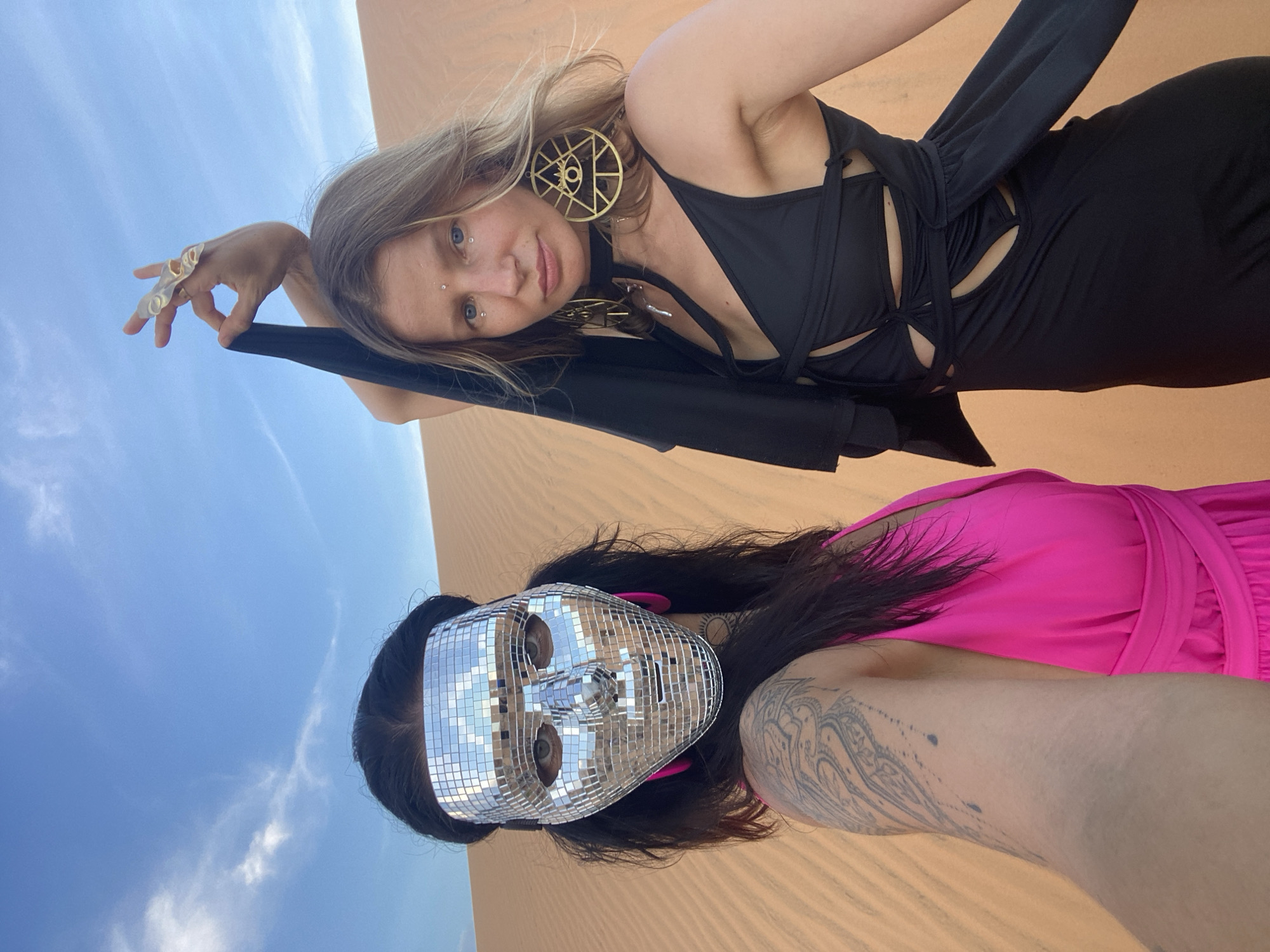 Two people are pictured in a desert landscape under a blue sky with sparse clouds. The person in the foreground is wearing a bright pink dress and a mirrorball mask that covers their face, revealing only their eyes. They have dark hair and a tattoo on their arm. The second person, in the background, has blonde hair and is wearing a black dress with cutouts and large, geometric gold earrings. They are looking at the camera and wearing rings on their fingers. The scene appears to be a posed photo shoot or a recreational activity in a desert environment. The lighting suggests it is daytime.