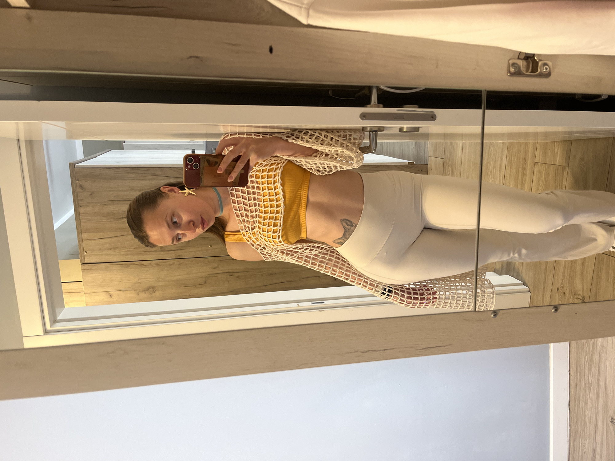 A woman is taking a selfie in a mirror. She is wearing a yellow top, white pants, and a white crochet cardigan. She has a blue choker and starfish earrings. She is holding a phone in her right hand. The background is a room with light wood-paneled walls and floors. The reflection in the mirror includes a vanity and cabinet. The location is Bečići, Montenegro.