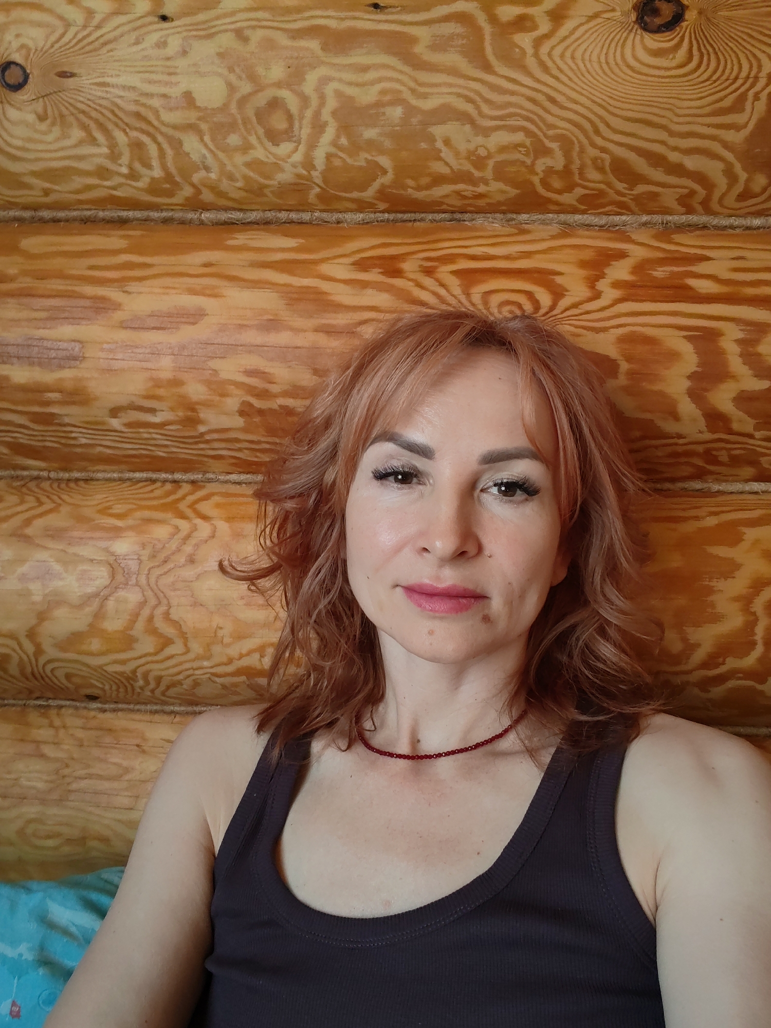 A woman with red hair and a brown tank top is sitting against a wooden log wall. She is wearing a red beaded necklace and has makeup on. She is facing the camera. The background suggests a rustic interior, possibly a log cabin or house constructed with logs. There is no information to determine the location is Старое Михайловское, Russia.