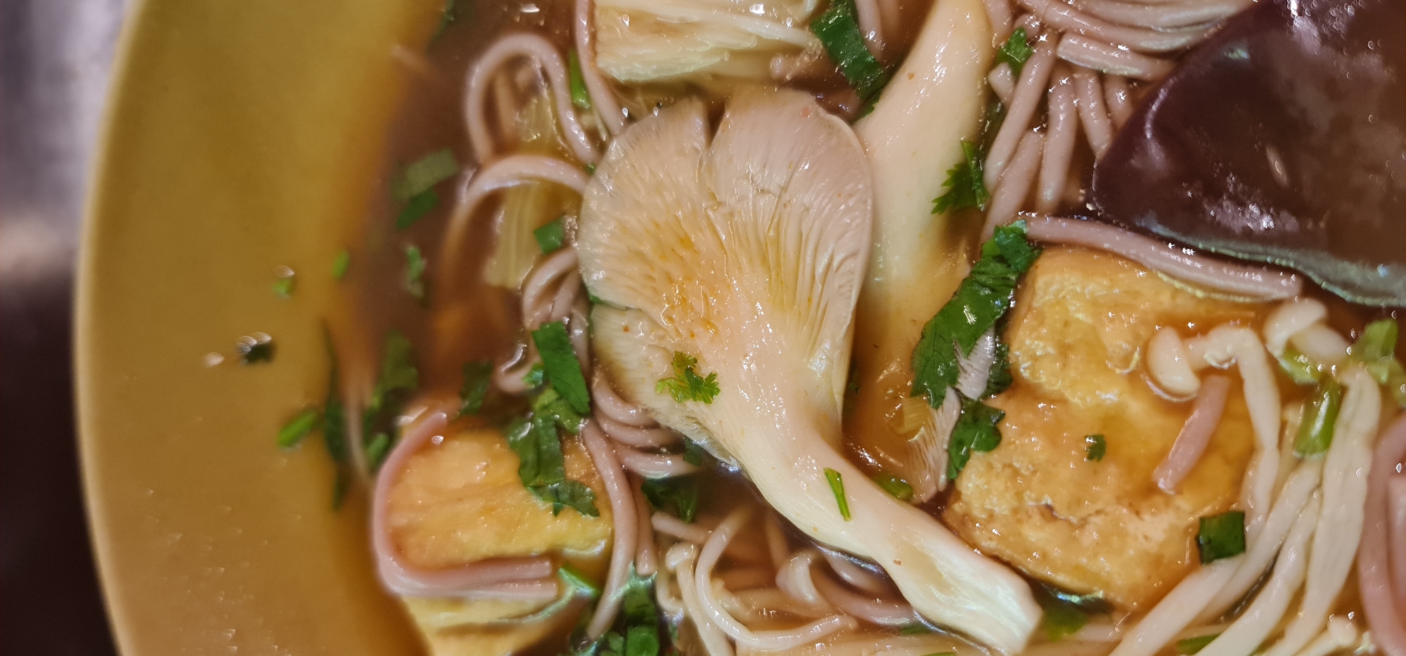 A close-up shot reveals a bowl of Vietnamese noodle soup, potentially located in Đà Nẵng, Vietnam. The soup contains soba noodles, tofu, various types of mushrooms, and chopped cilantro. The broth appears clear, and the ingredients suggest a flavorful and balanced meal. The bowl itself is a yellowish color.