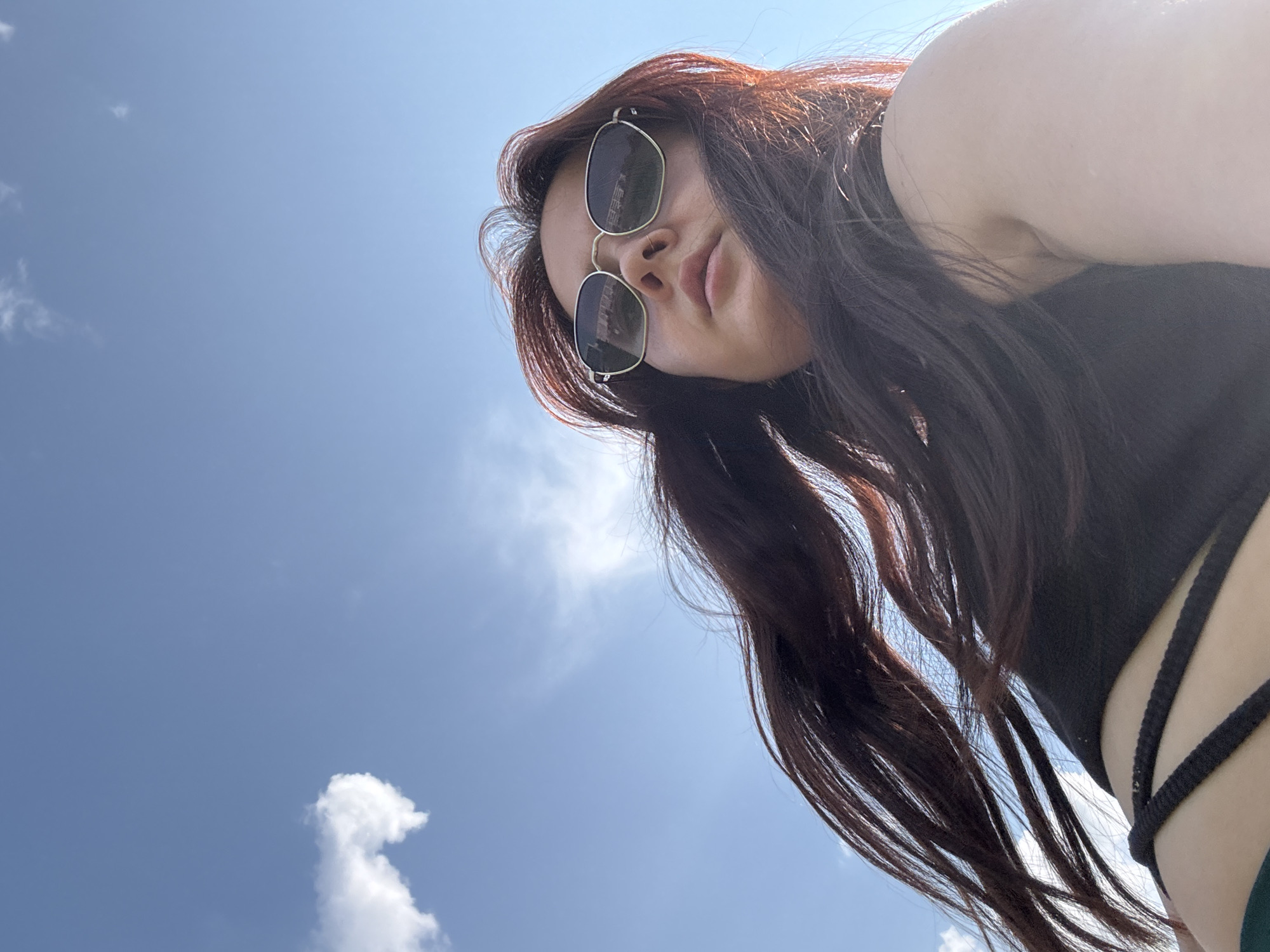 A low-angle, close-up outdoor photograph shows a young woman wearing sunglasses and a black top against a bright blue sky with scattered clouds. Her long, dark hair is blowing in the wind, framing her face. The woman is looking upwards, with her mouth slightly open. The lighting suggests it is daytime. No specific location within Naberezhnye Chelny, Russia, can be determined from the image alone.