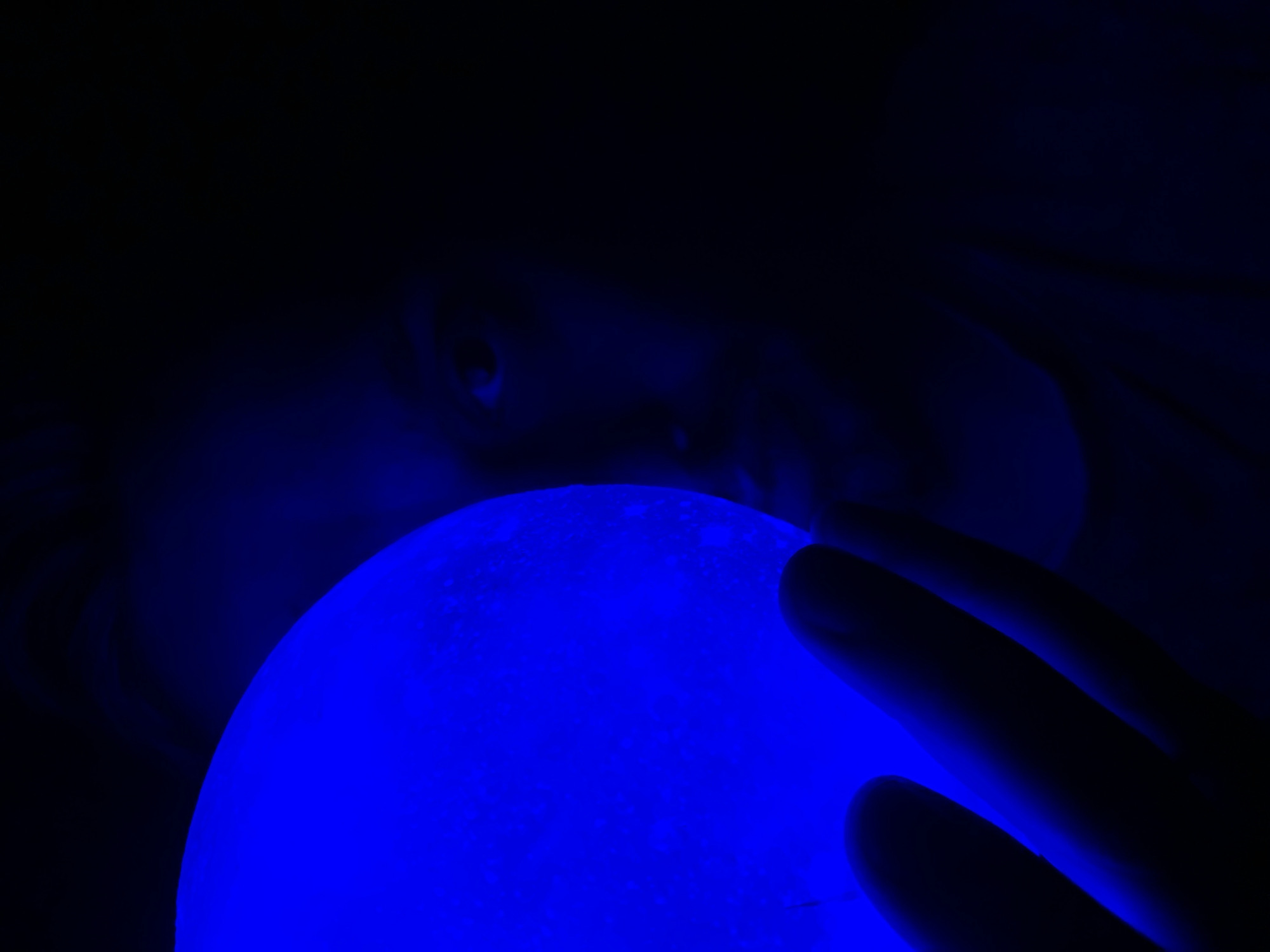 A close-up, low-light image features a person partially visible in the background, looking down at a bright blue, spherical object held by their hand. The person's eyes and nose are discernible, but their features are obscured by the darkness. The blue sphere is illuminated and textured, suggesting a moon-like surface. A hand is partly visible on the right, appearing to hold or touch the sphere. The background is predominantly black. The location in the city of Chaikovsky, Russia cannot be determined from the image alone.