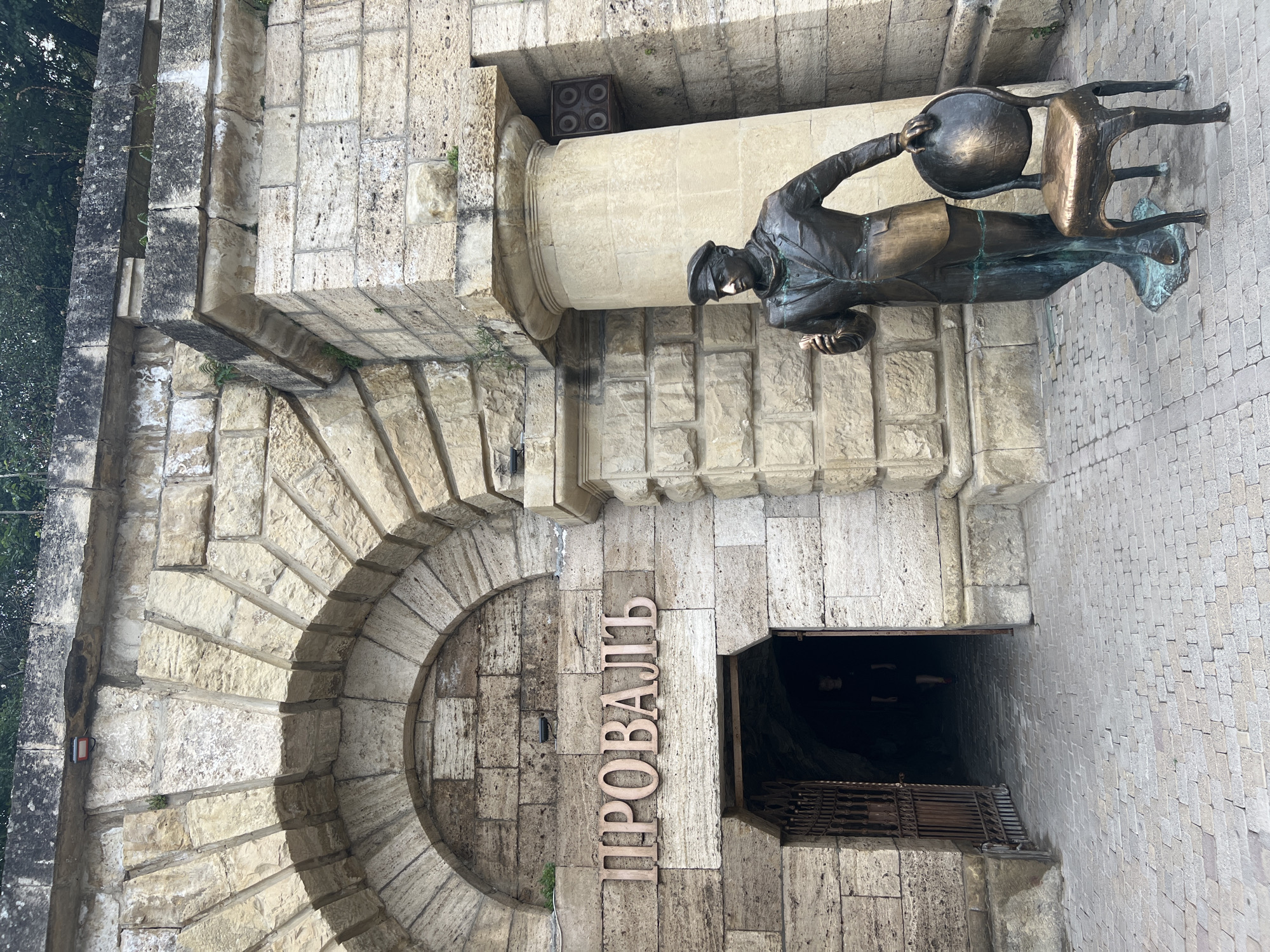 The image depicts the entrance to the Proval, a sulfurous lake in Pyatigorsk, Russia. A bronze statue of a man in a suit and cap stands to the right of the entrance, holding an object in his right hand and resting his left hand on the back of a bronze chair. The entrance itself is an arched doorway with weathered stone walls and a wrought-iron gate. The word "ПРОВАЛЬ" (Proval) is inscribed in large wooden letters above the entrance. The ground is paved with cobblestones. In the background, a person is visible inside the dark entrance. The architecture suggests an older, historic structure.